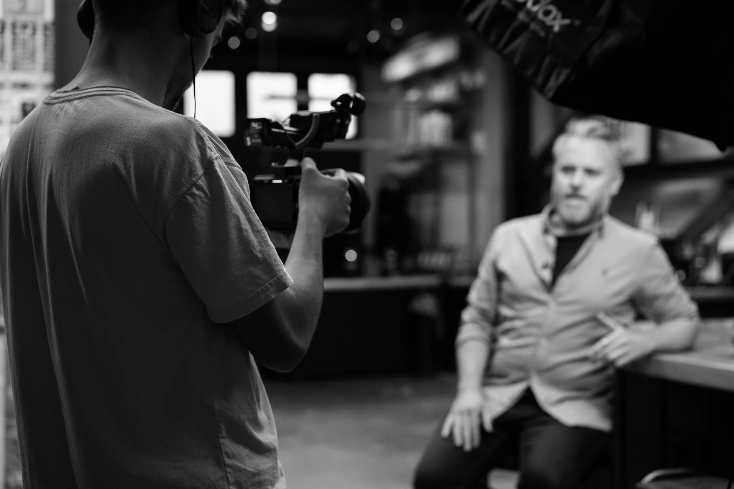 A person holding a camera and filming another person sitting on a chair in an indoor setting with shelves in the background.