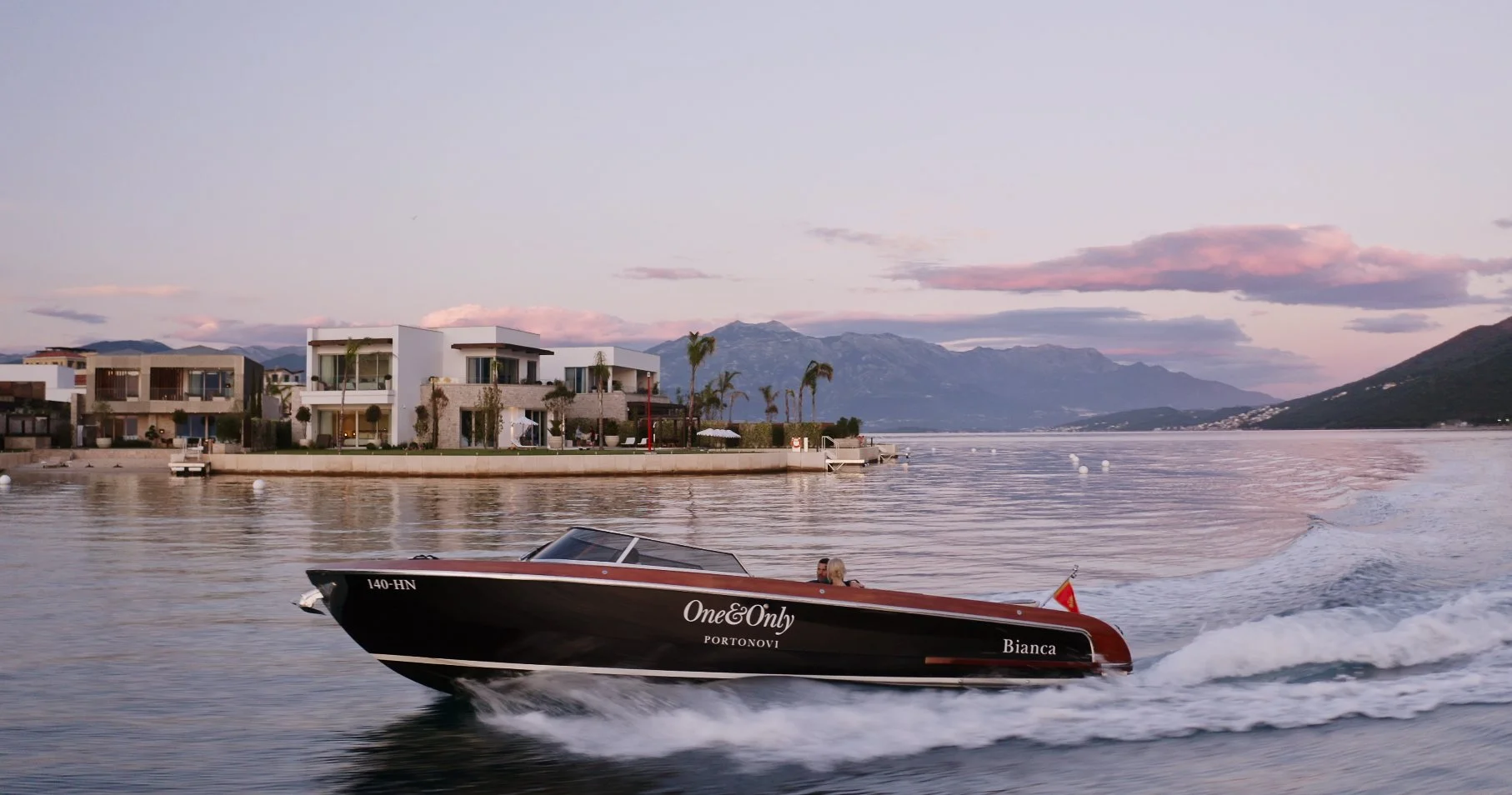 A speedboat named 'Bianca' moving quickly across a lake or river, with modern waterfront houses and mountains in the background during sunset.