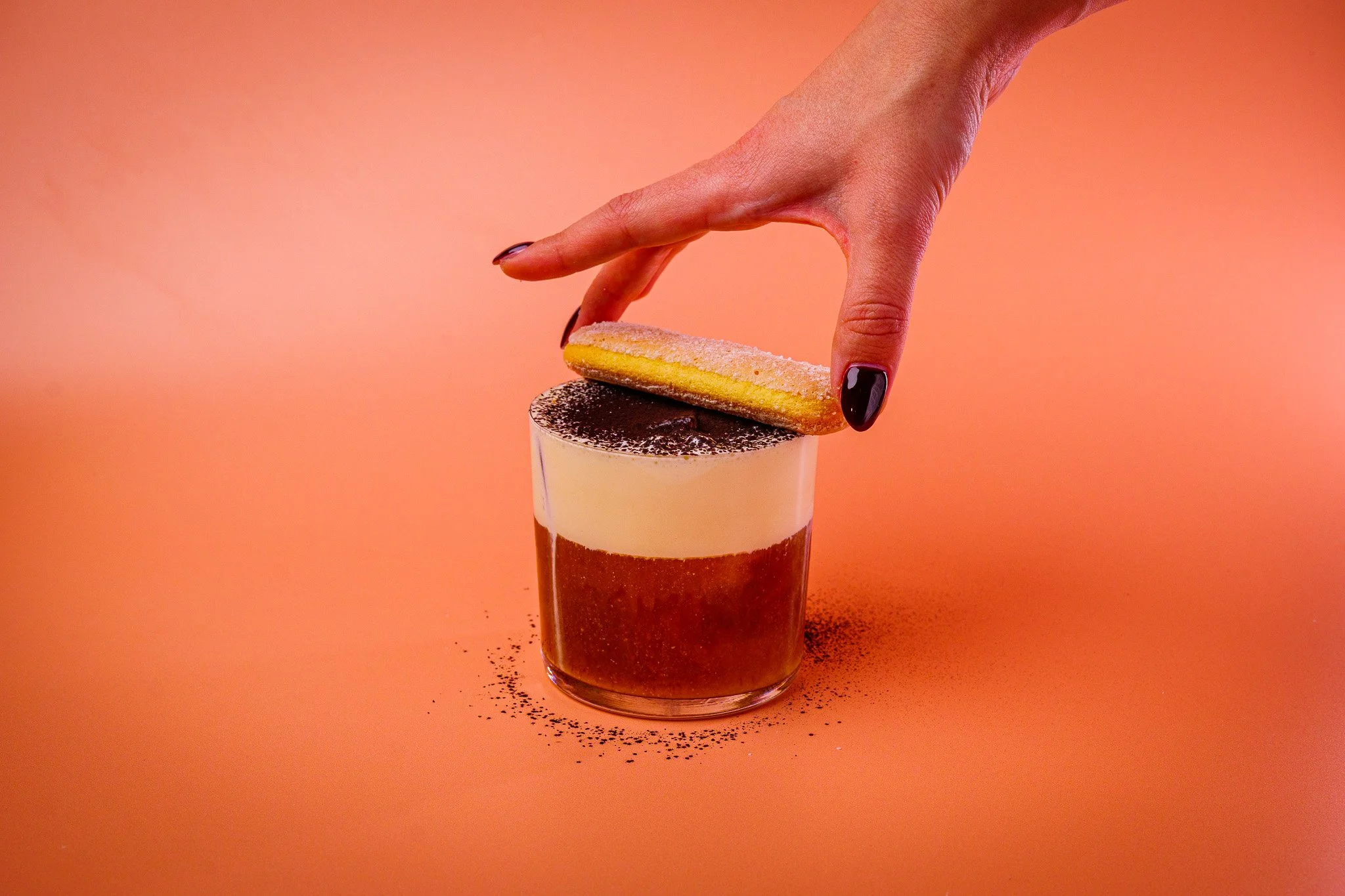 Layered coffee drink in a glass topped with cocoa powder, a lady's hand with dark-colored nail polish holding a yellow and pink almond cookie over the drink against a peach-colored background.
