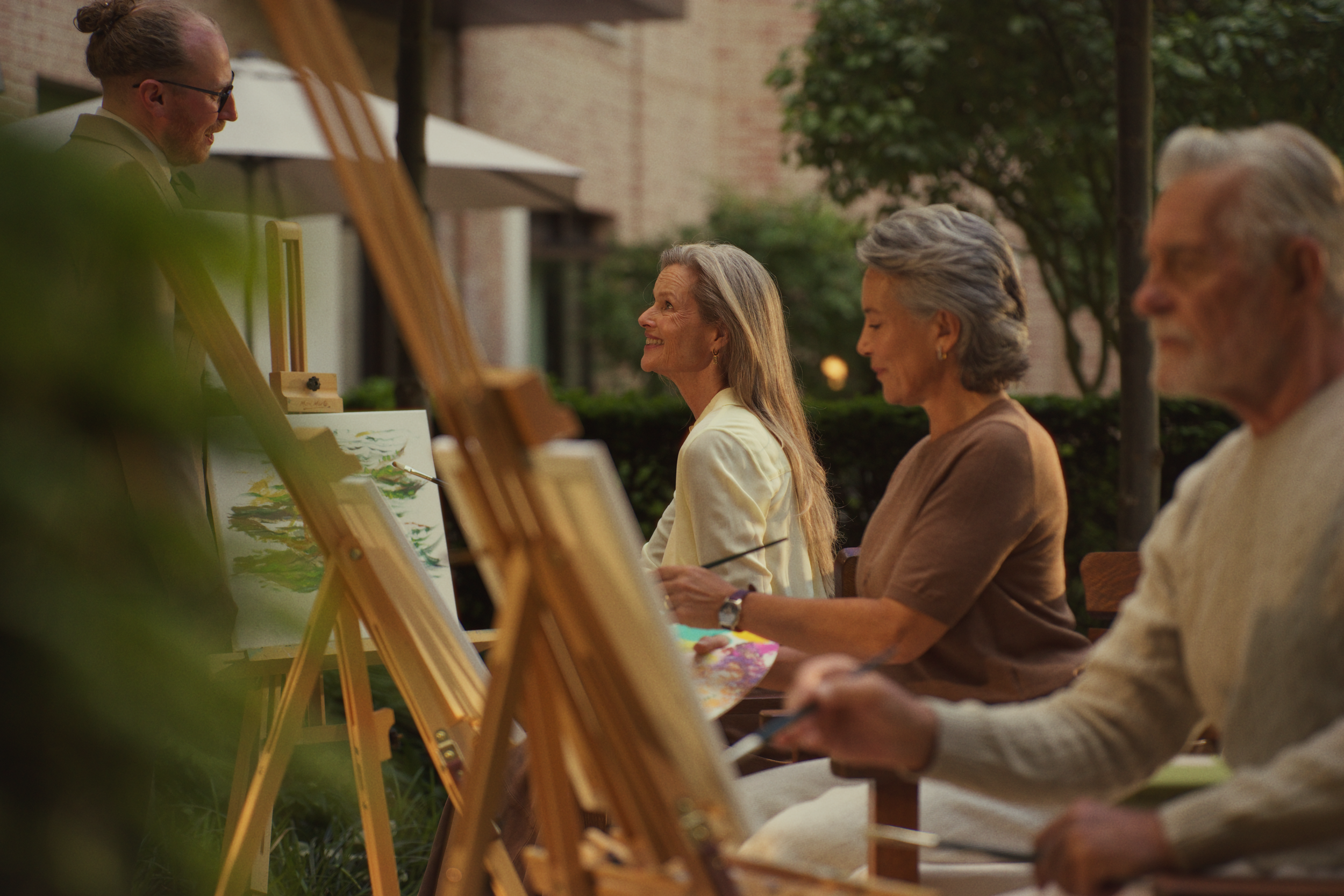 A group of elderly people painting on canvases set up on easels outdoors, with a man standing and talking to the women, in a garden or park setting.