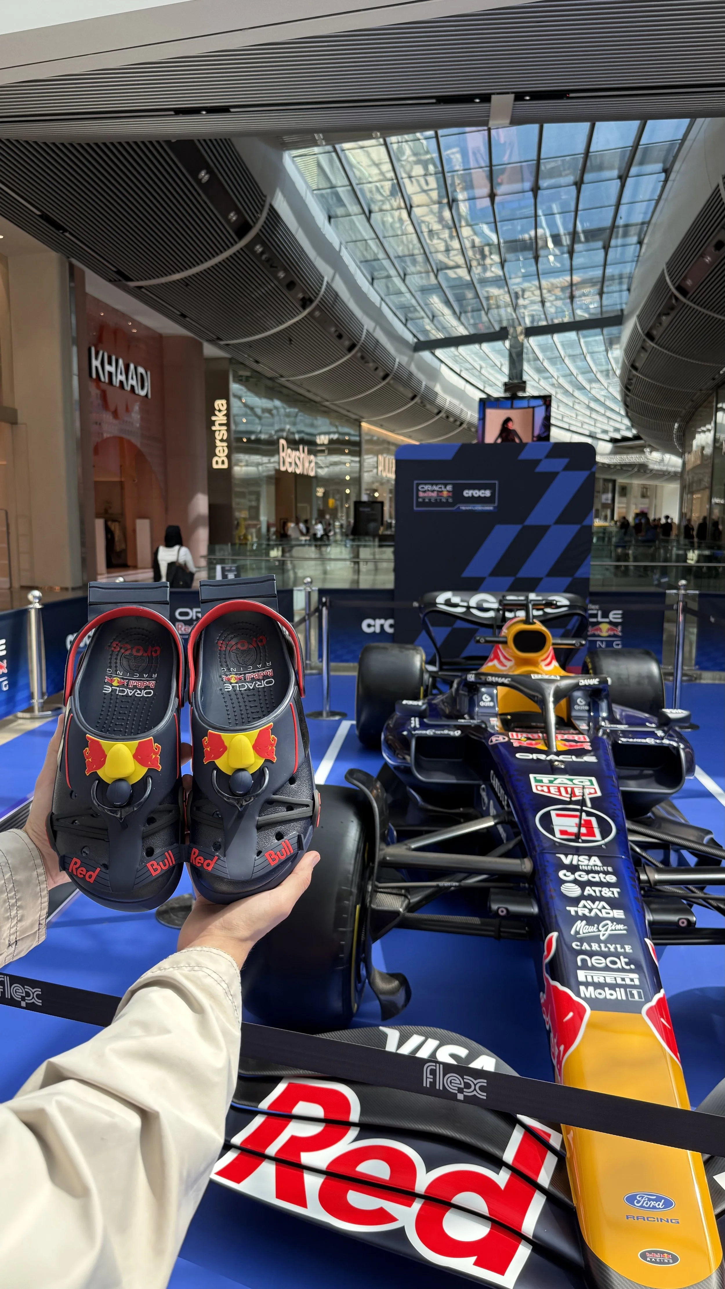 A pair of Red Bull Racing Formula 1 racing shoes held in front of a Red Bull Racing Formula 1 car, with a yellow and black Ford racing car in the background inside a modern shopping mall.