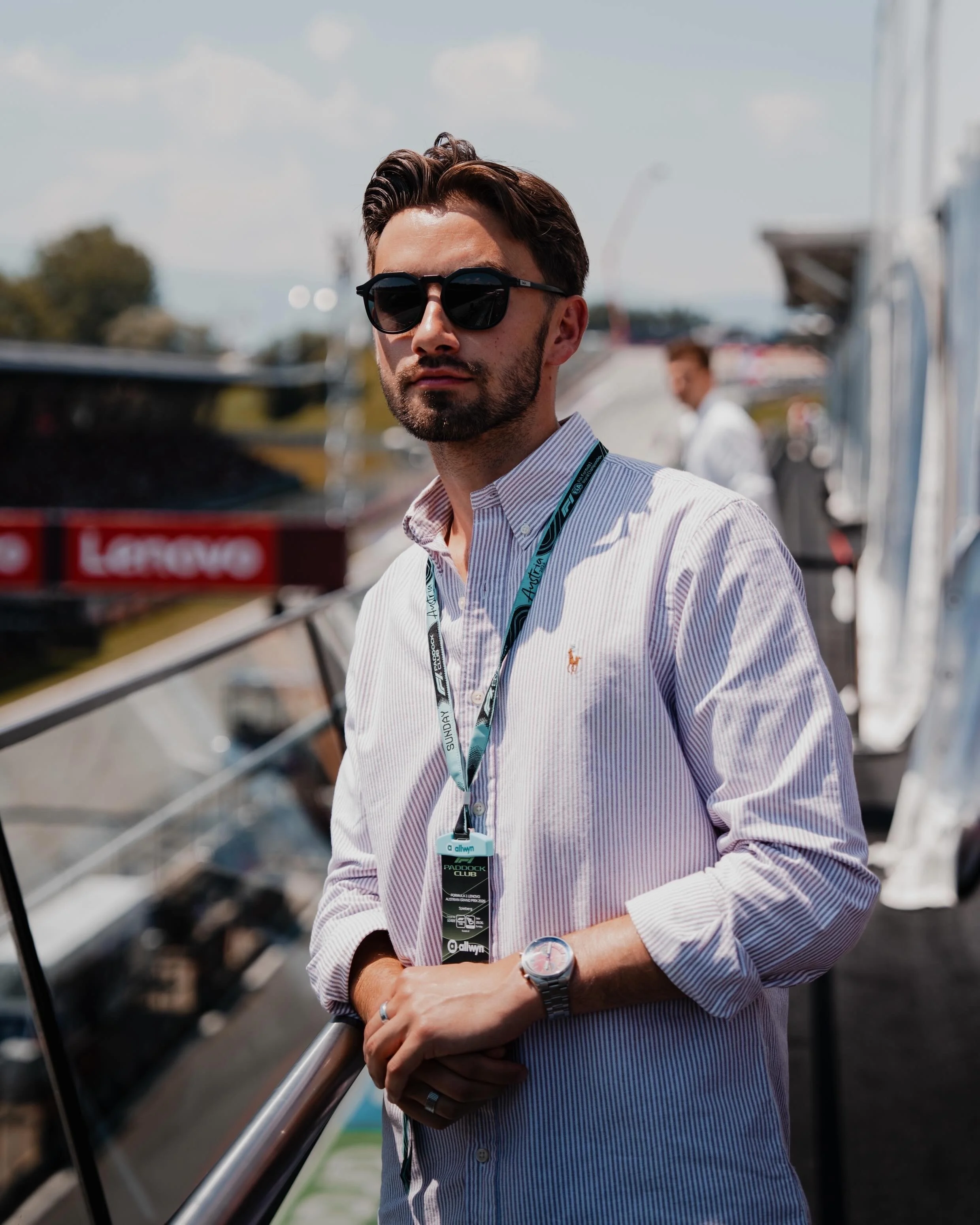 A man with dark hair, beard, and sunglasses wearing a striped button-down shirt and a lanyard, standing outdoors on a bright day, with boats and a marina in the background.