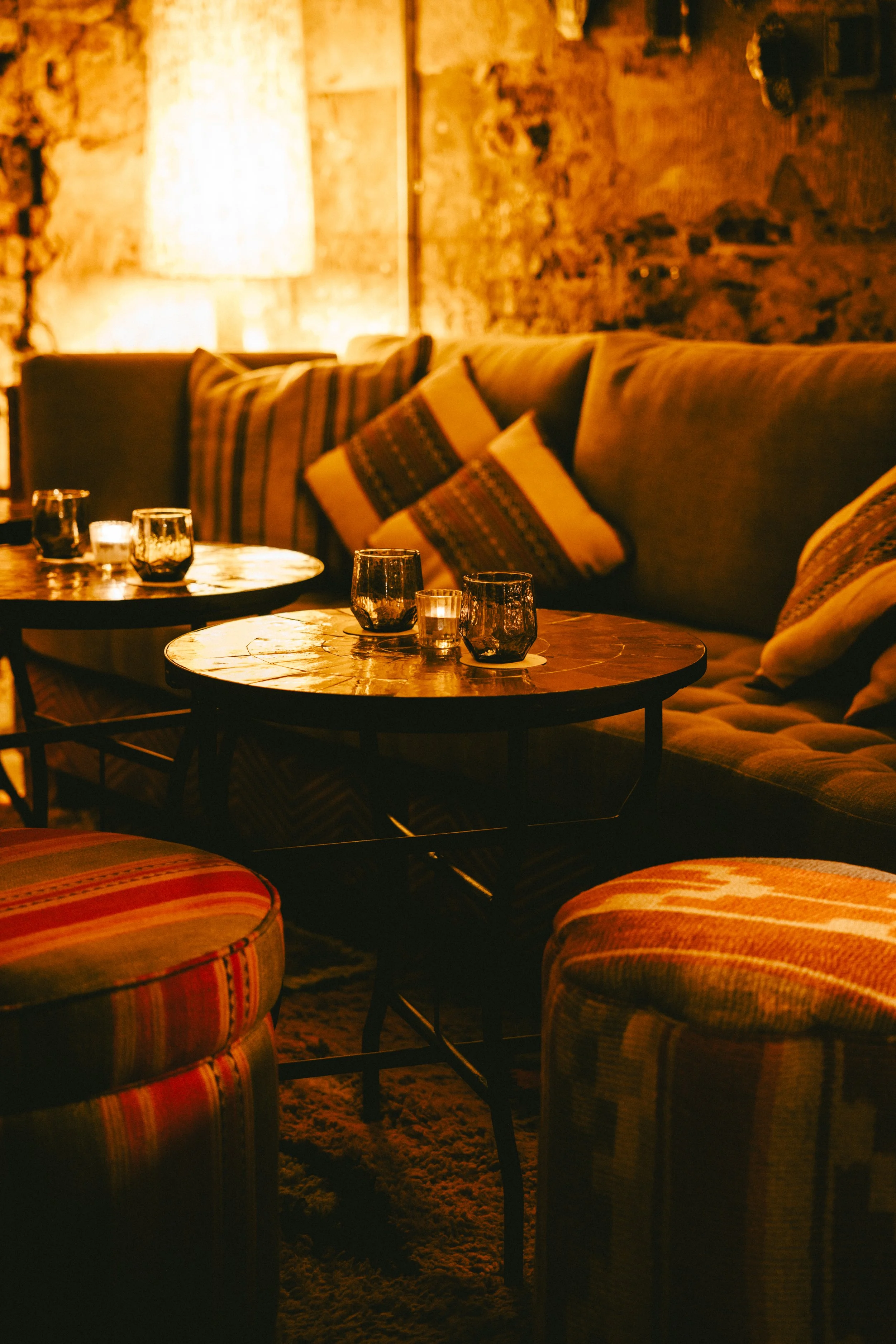 A cozy living room with a brown leather sofa adorned with colorful patterned cushions, two wooden coffee tables with glass candle holders, a wooden side table with a lamp, exposed brick wall, and warm ambient lighting.