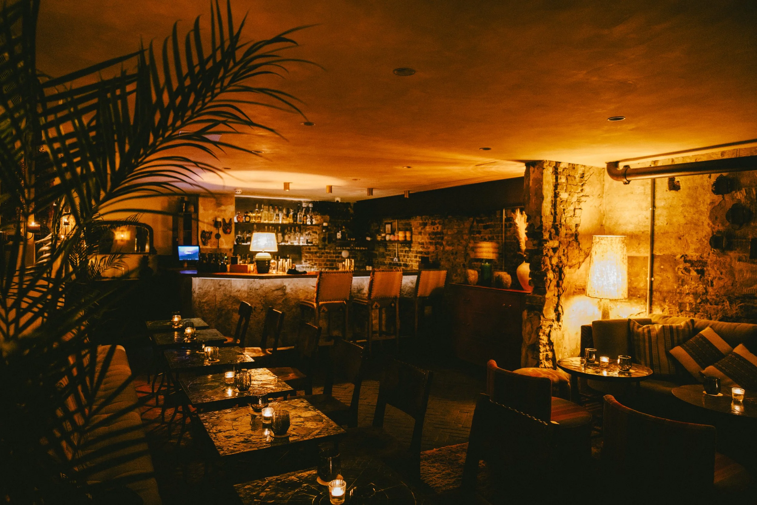 Dimly-lit bar area with a curved brick wall, shelves stocked with bottles, wooden bar stools, tables with lit candles, and warm orange lighting creating a cozy atmosphere.