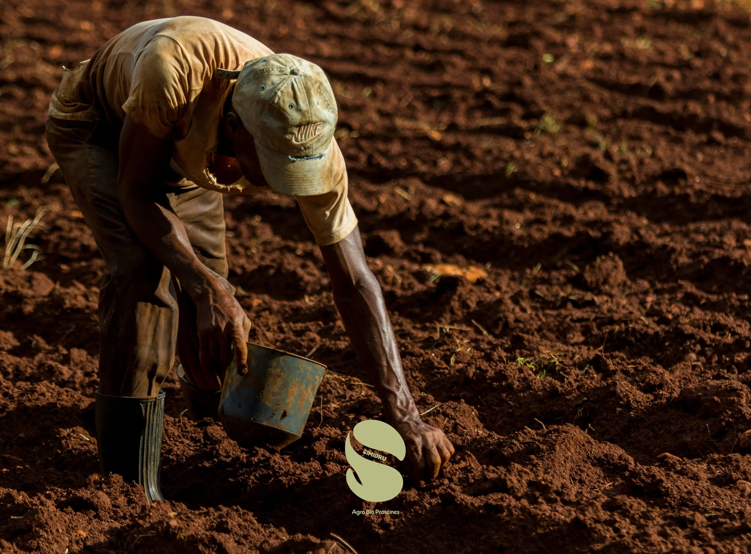 Un homme travaille dans un champ de terre rouge, portant un t-shirt beige, un chapeau de baseball, et une botte en caoutchouc, avec une bêche à la main, dans un environnement agricole.