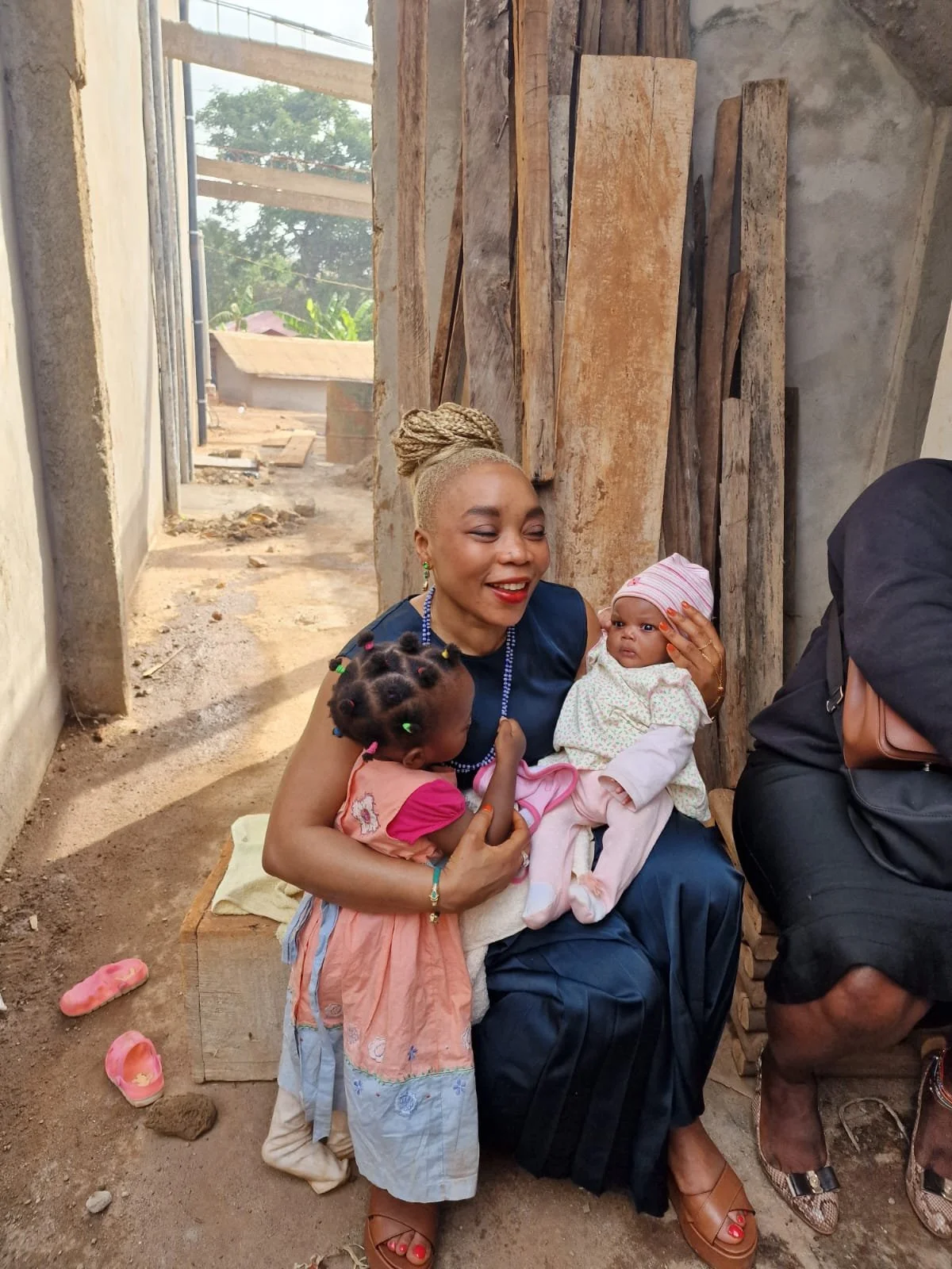 Une femme souriante tient deux enfants dans un environnement en construction avec du bois brut en arrière-plan.