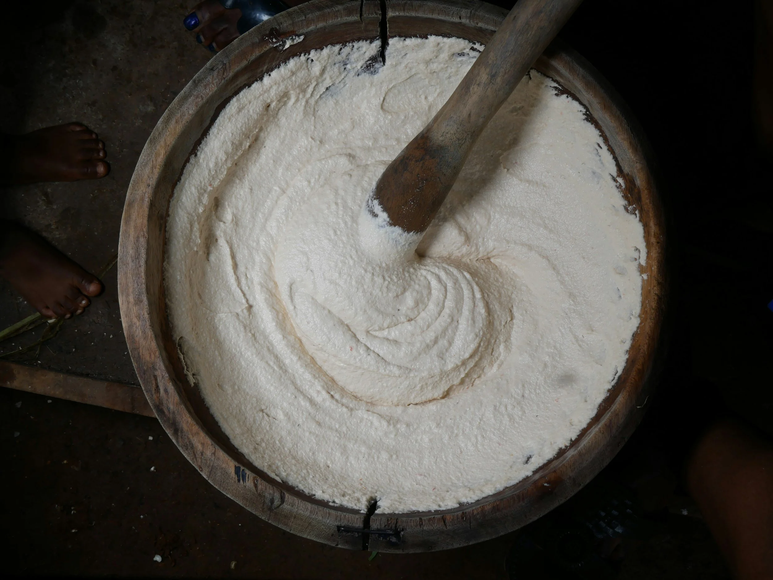 Préparation de lait de soja dans un grand récipient en bois, avec un pilon en bois et un bec verseur en métal. Deux personnes sont visibles, une à gauche avec les pieds nus et une autre à droite avec des chaussures en plastique.