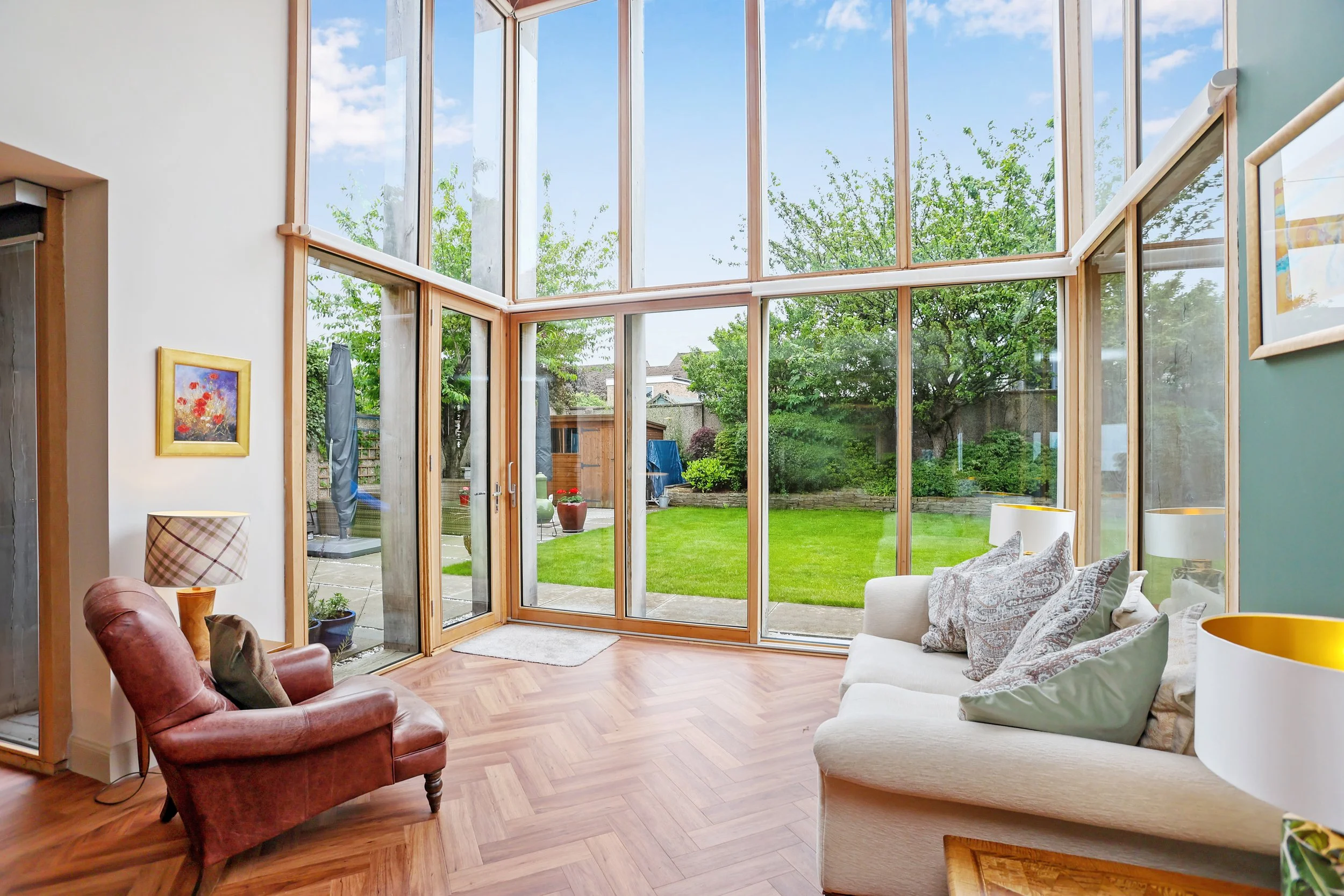 A bright living room with large glass windows and doors revealing a green backyard with trees, potted plants, and blue sky. The room has a wooden floor and is furnished with a white sofa, an armchair, and lamps.