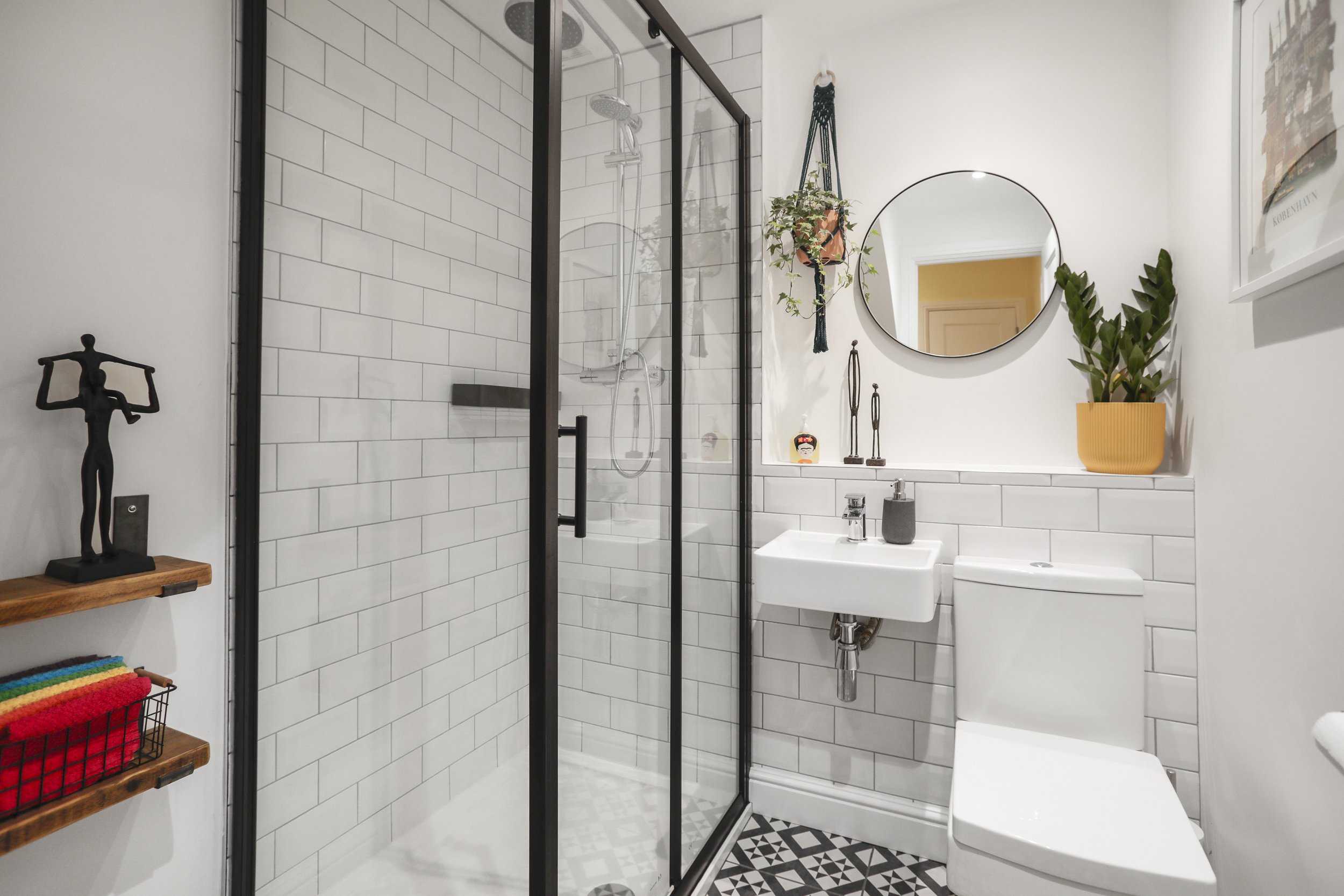 A bathroom with a glass-enclosed shower area that has white subway tiles and a rainfall showerhead. There is a round mirror above a white sink, and a toilet is adjacent. Decor includes a potted plant, hanging plants, and a figurine, with shelves hold