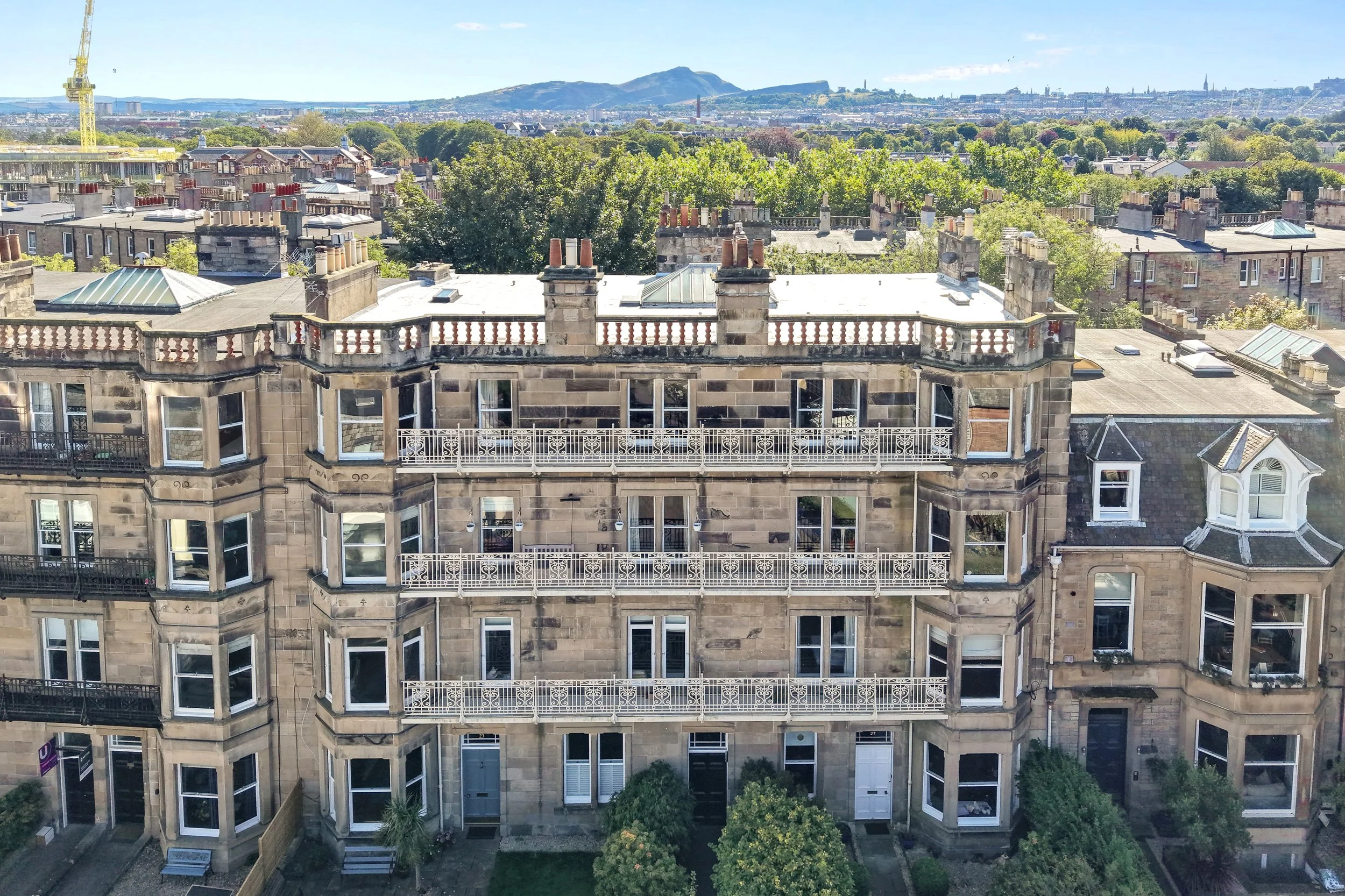 A high-rise building with stone exterior walls, large bay windows, and white balconies. The rooftop features multiple chimneys, some with red clay pots, and several glass skylights. There are trees and a cityscape with mountains in the background.
