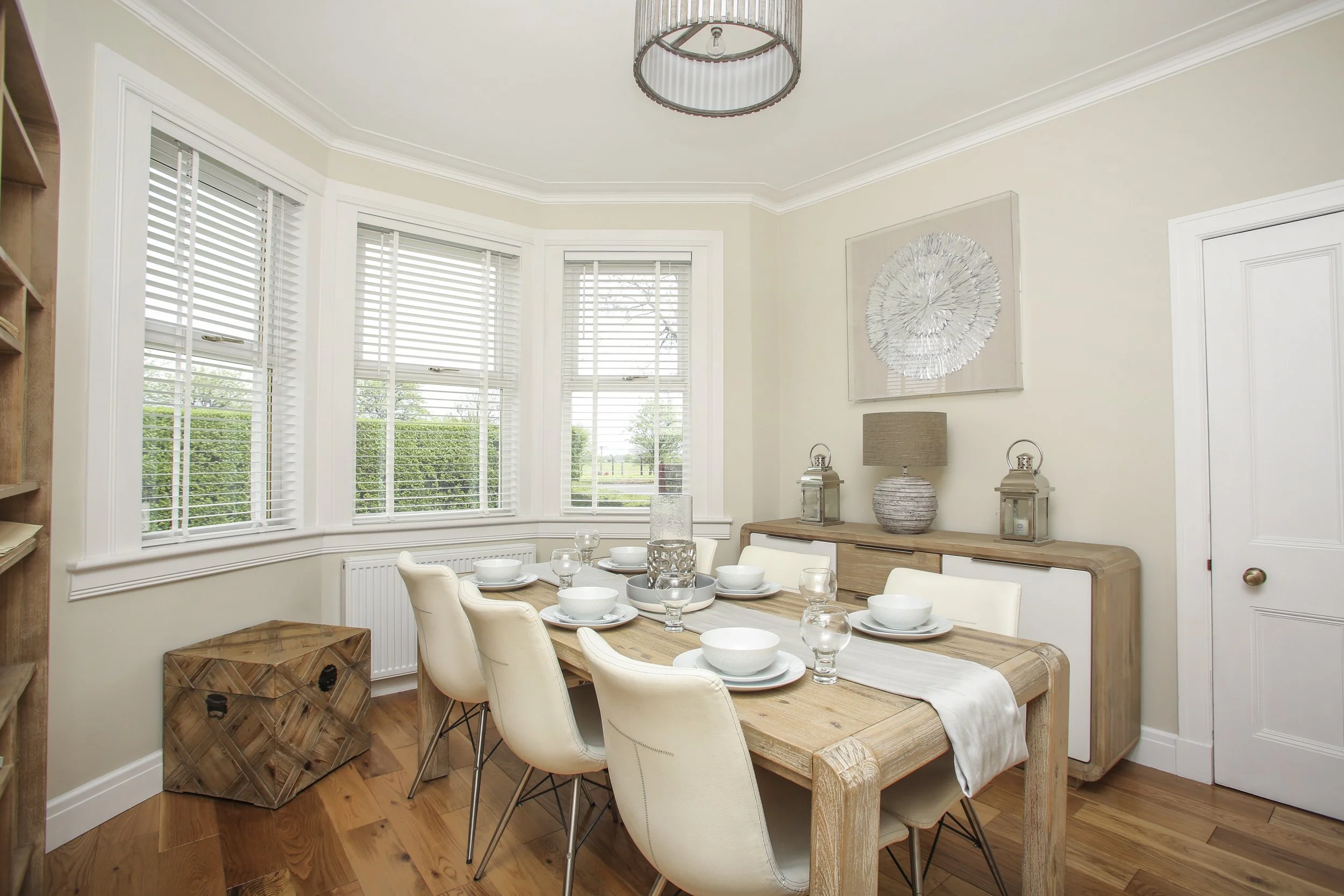 A dining room with a wooden table set with white dishes, glasses, and a table runner, surrounded by cream-colored chairs, with a large window with blinds, a sideboard with decorative lamps, and artwork on the wall.
