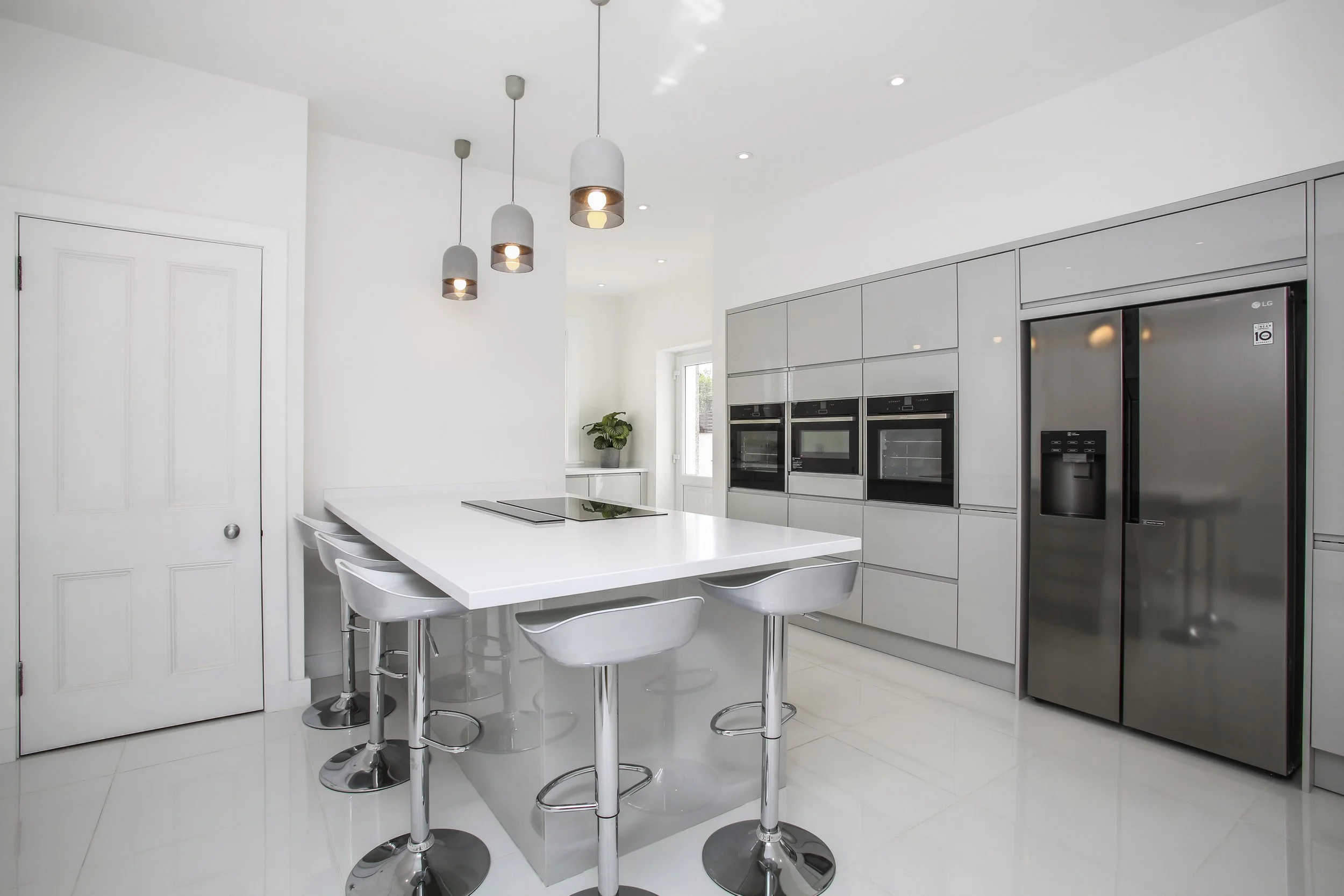 Modern white kitchen with a breakfast bar, gray cabinetry, built-in appliances, a stainless steel refrigerator, pendant lights, and a small window with a potted plant.
