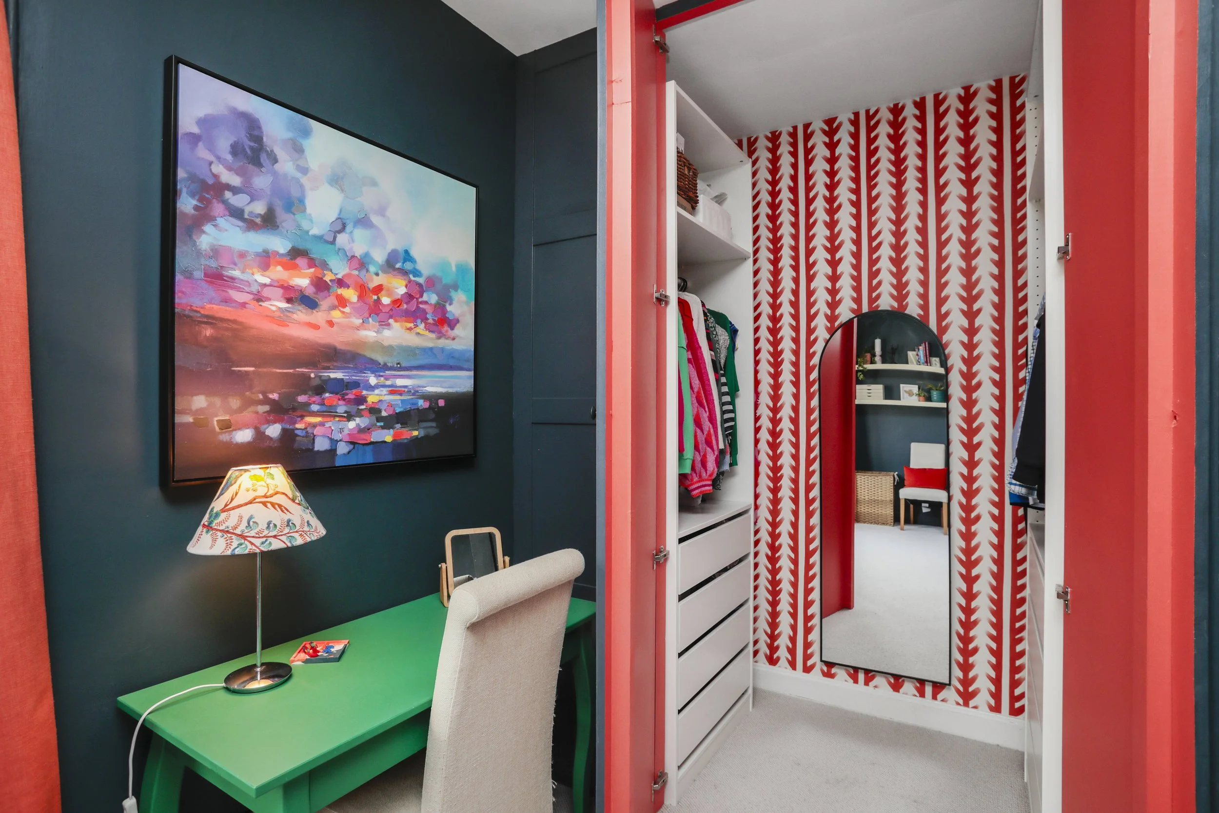 A closet with red and white patterned wallpaper, white shelving, and clothing hanging inside. Adjacent to the closet, part of a room with dark teal walls, a colorful abstract painting, a green desk, and a beige chair.