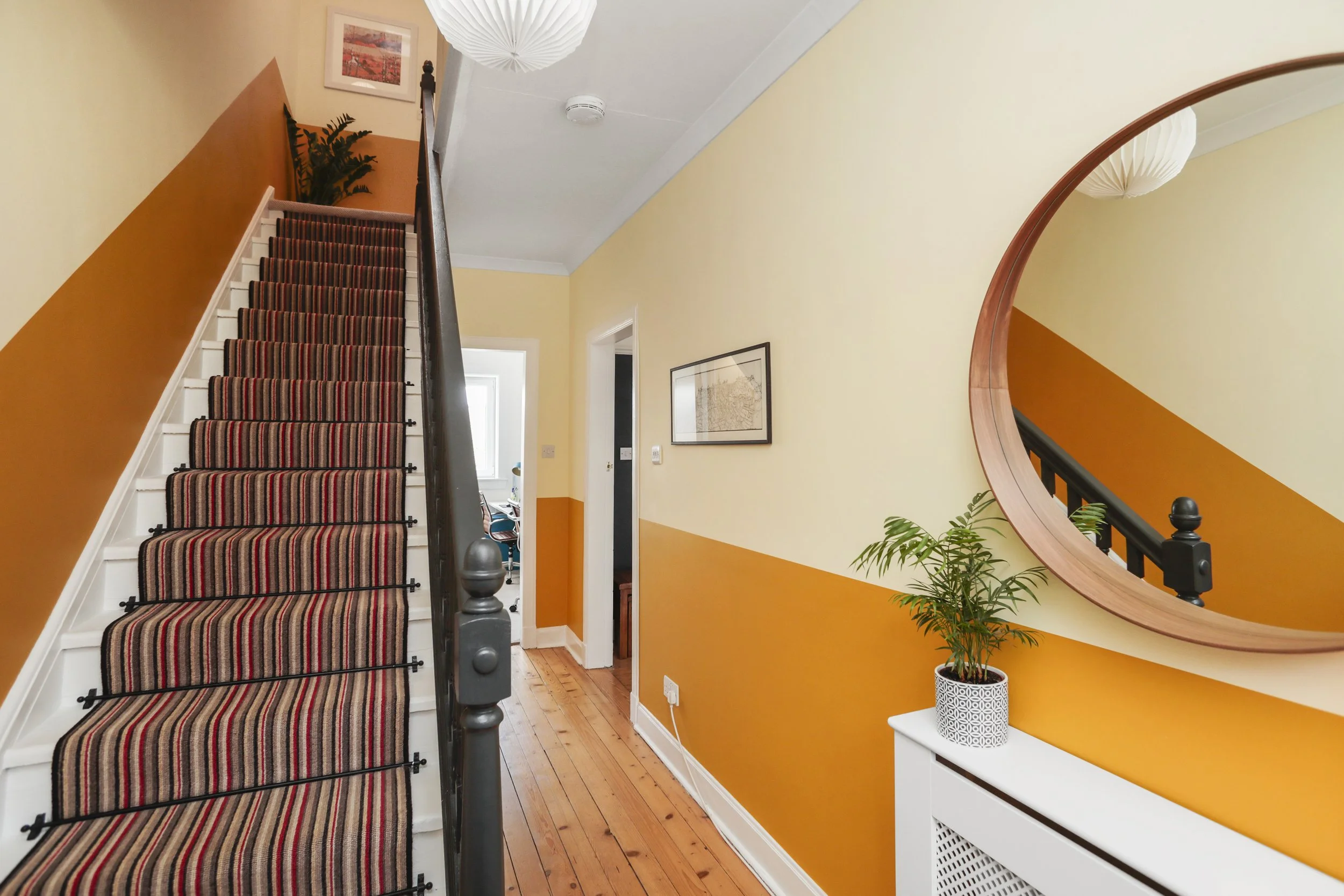 Interior view of a staircase with striped carpet, yellow and orange walls, a large round mirror on the right, potted plant on a white cabinet, framed picture on the wall, ceiling light, smoke detector, and doorways leading to other rooms.