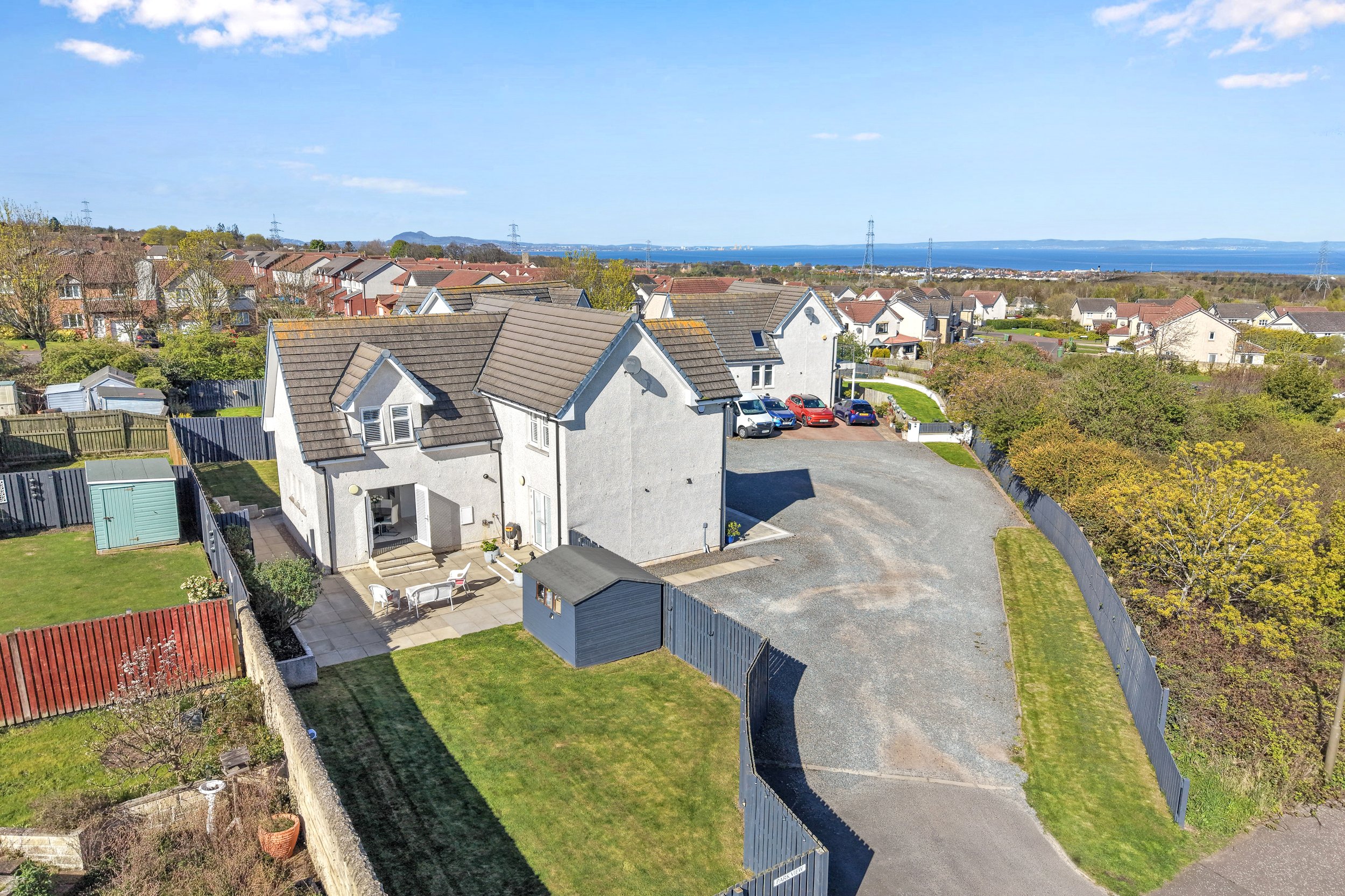 Aerial view of a residential neighborhood near a body of water, showing houses, parked cars, trees, and a clear blue sky.