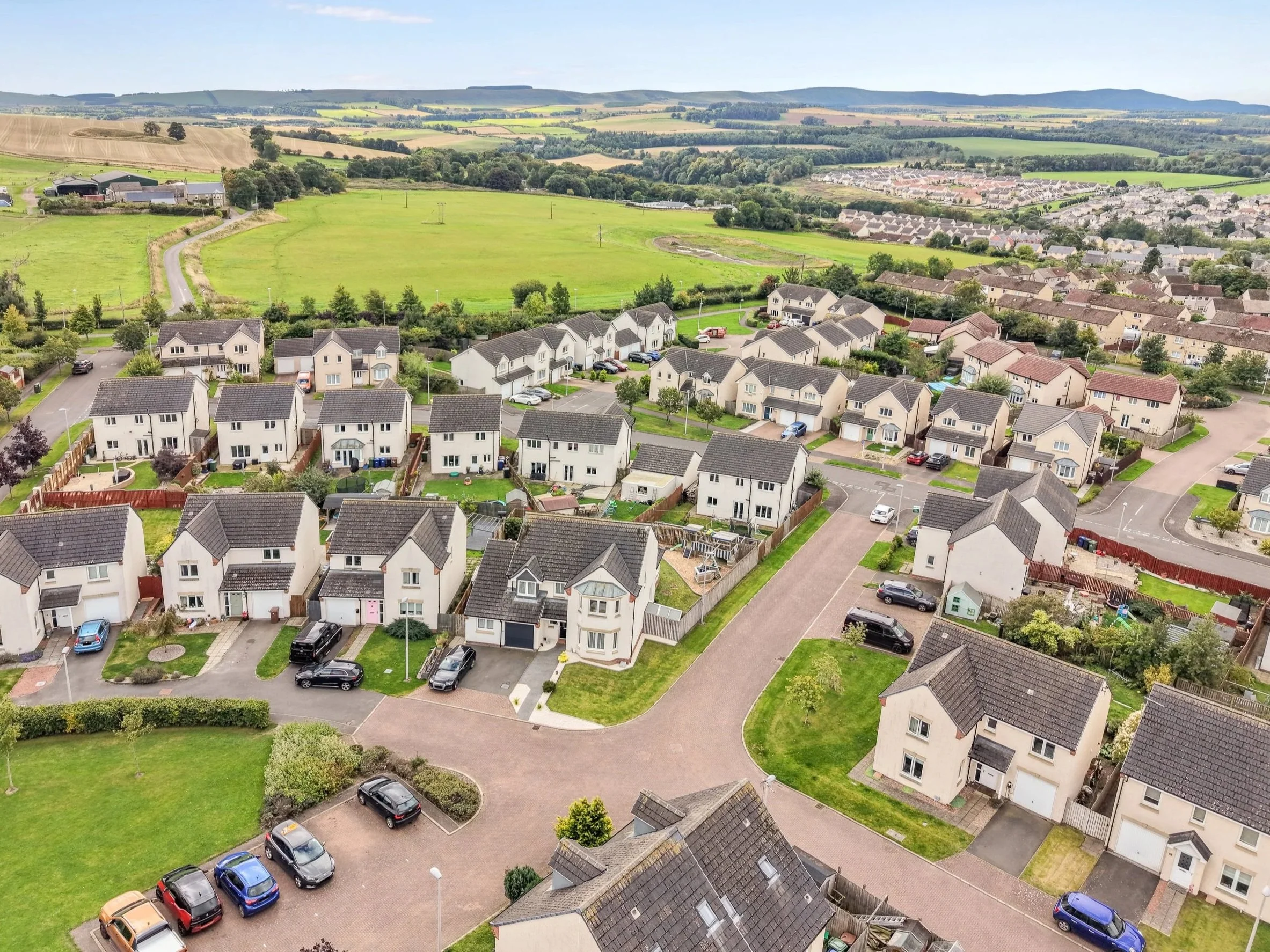 Aerial view of a suburban neighborhood with houses, streets, and green lawns, overlooking agricultural fields and rolling hills in the distance under a blue sky.