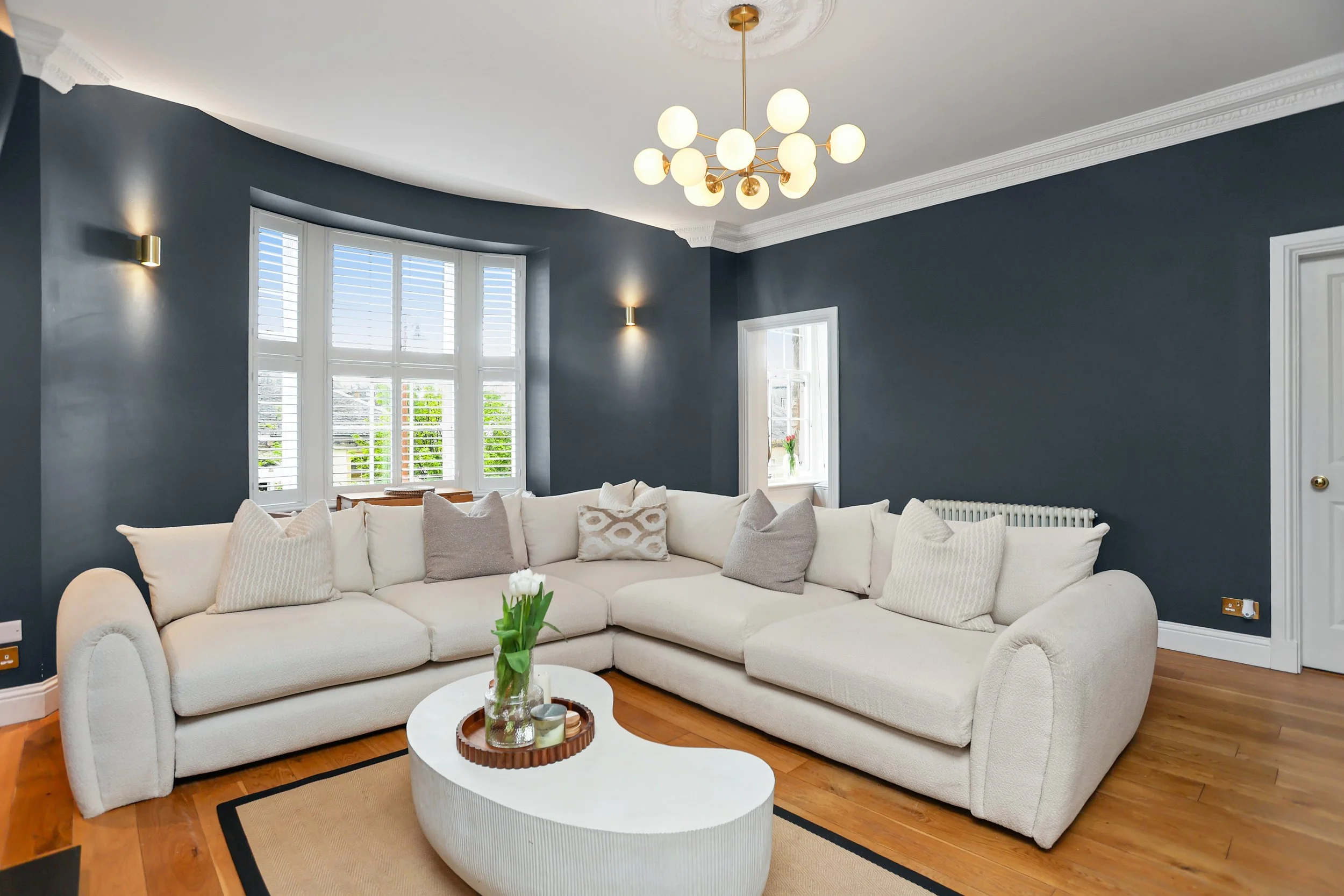 Living room with a large white sofa, beige and patterned pillows, a round coffee table with a vase of tulips, dark blue walls, wooden flooring, large windows with white blinds, and a modern chandelier and wall lights.