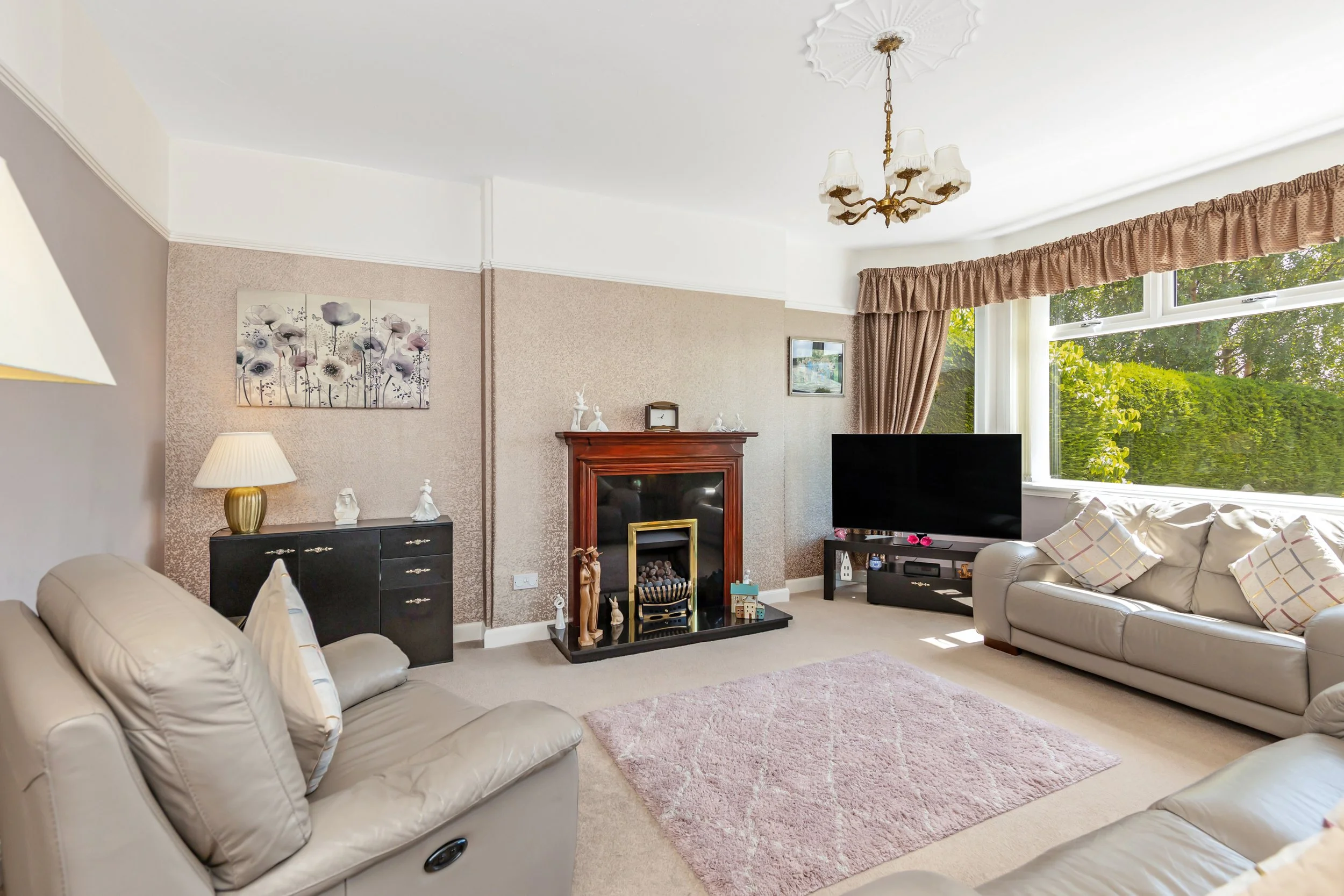 Living room with two beige sofas, a pink rug, a black cabinet, a television, a fireplace with decorative items, a framed picture, a window with curtains, and a ceiling chandelier.
