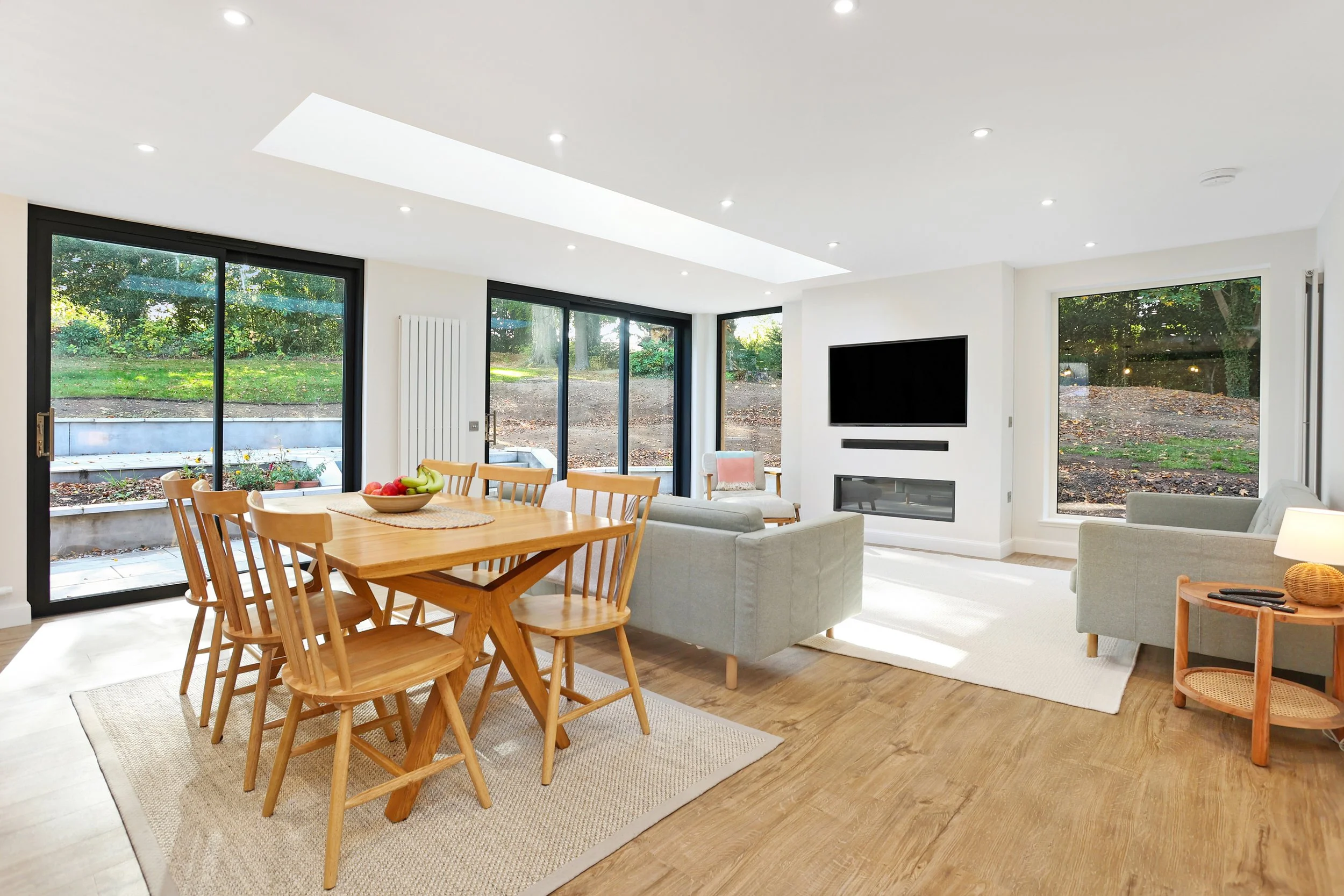A spacious, bright living and dining room with large windows showing an outdoor garden. Features include a wooden dining table with chairs, a gray sofa, a TV mounted on the wall, and a patterned rug on hardwood floors.