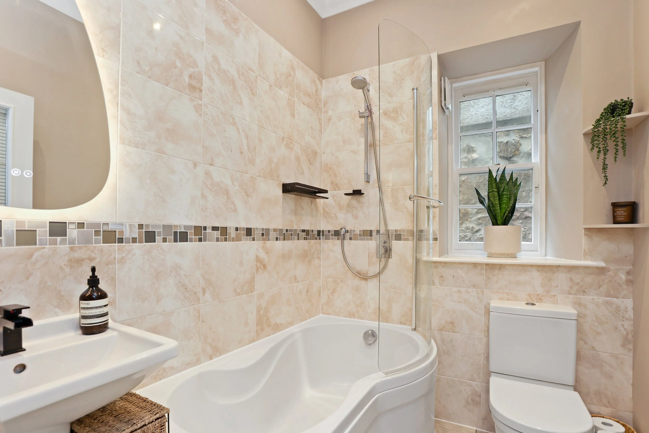 Bathroom with beige tiled walls, a bathtub with a curved edge, a wall-mounted showerhead, a small white sink with a soap dispenser, a white toilet, a window with frosted glass, and decorative plants on a shelf.