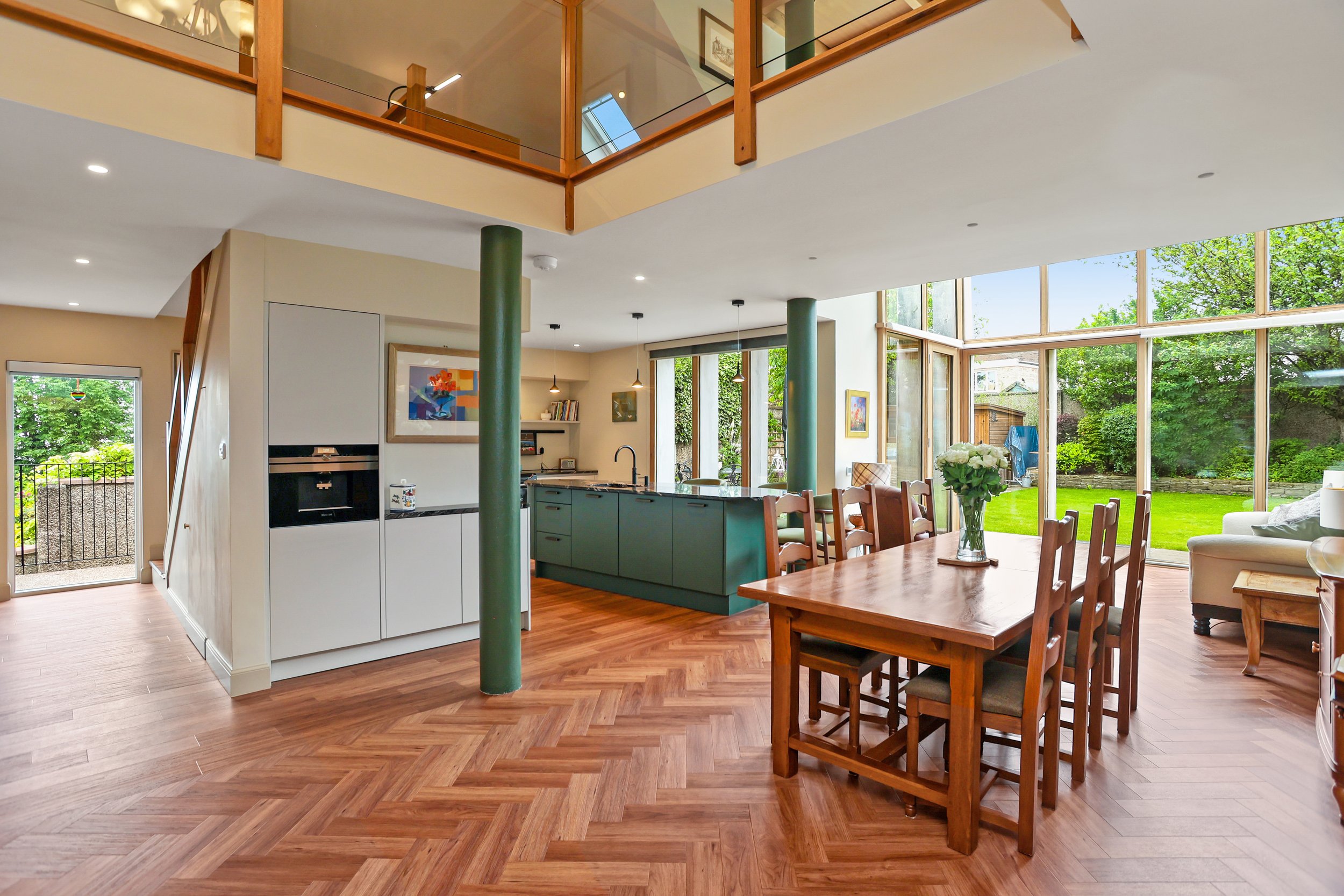 Open-concept living space with large windows, wooden flooring, a wooden dining table with chairs, a kitchen with a green island, and a view of a green backyard.