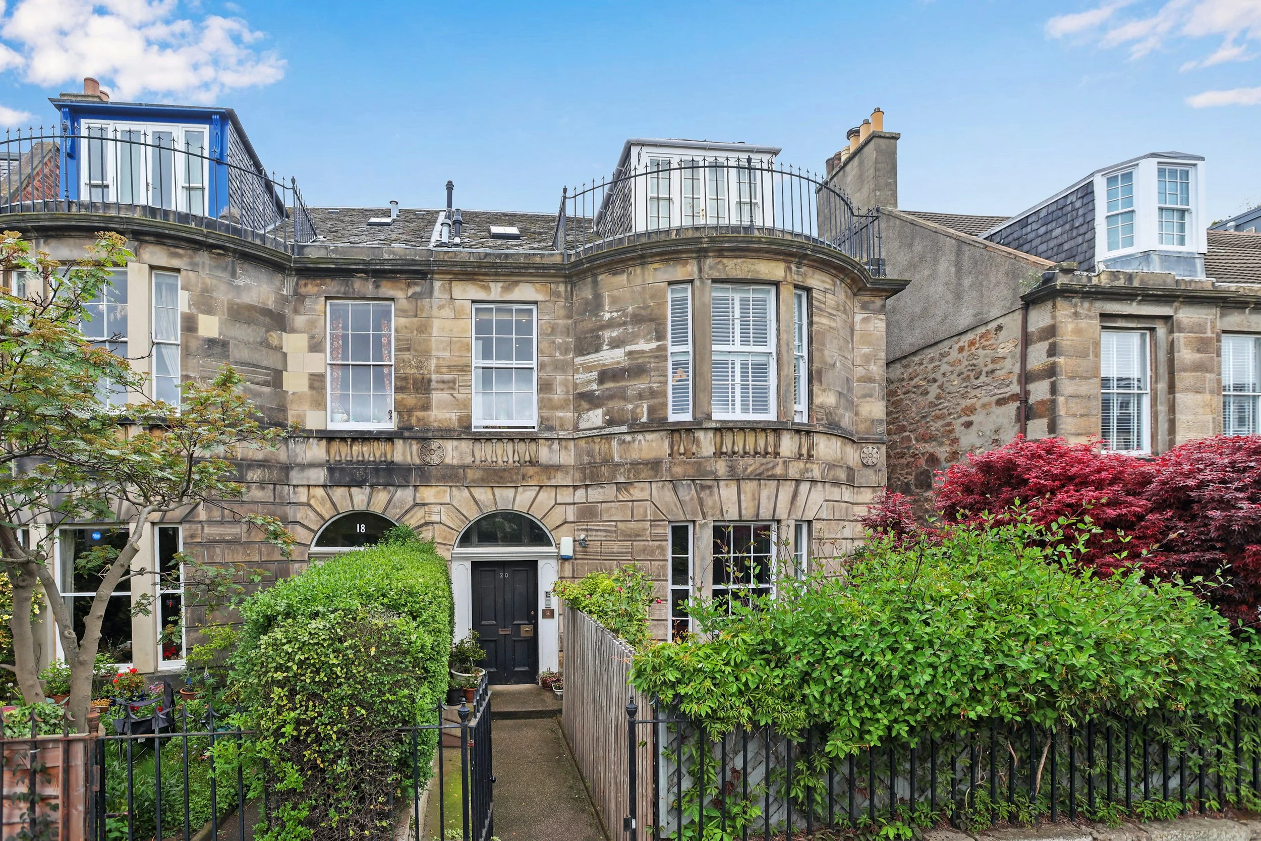 Front view of a historic stone townhouse with multiple windows and a garden at the entrance, along with surrounding greenery and trees.