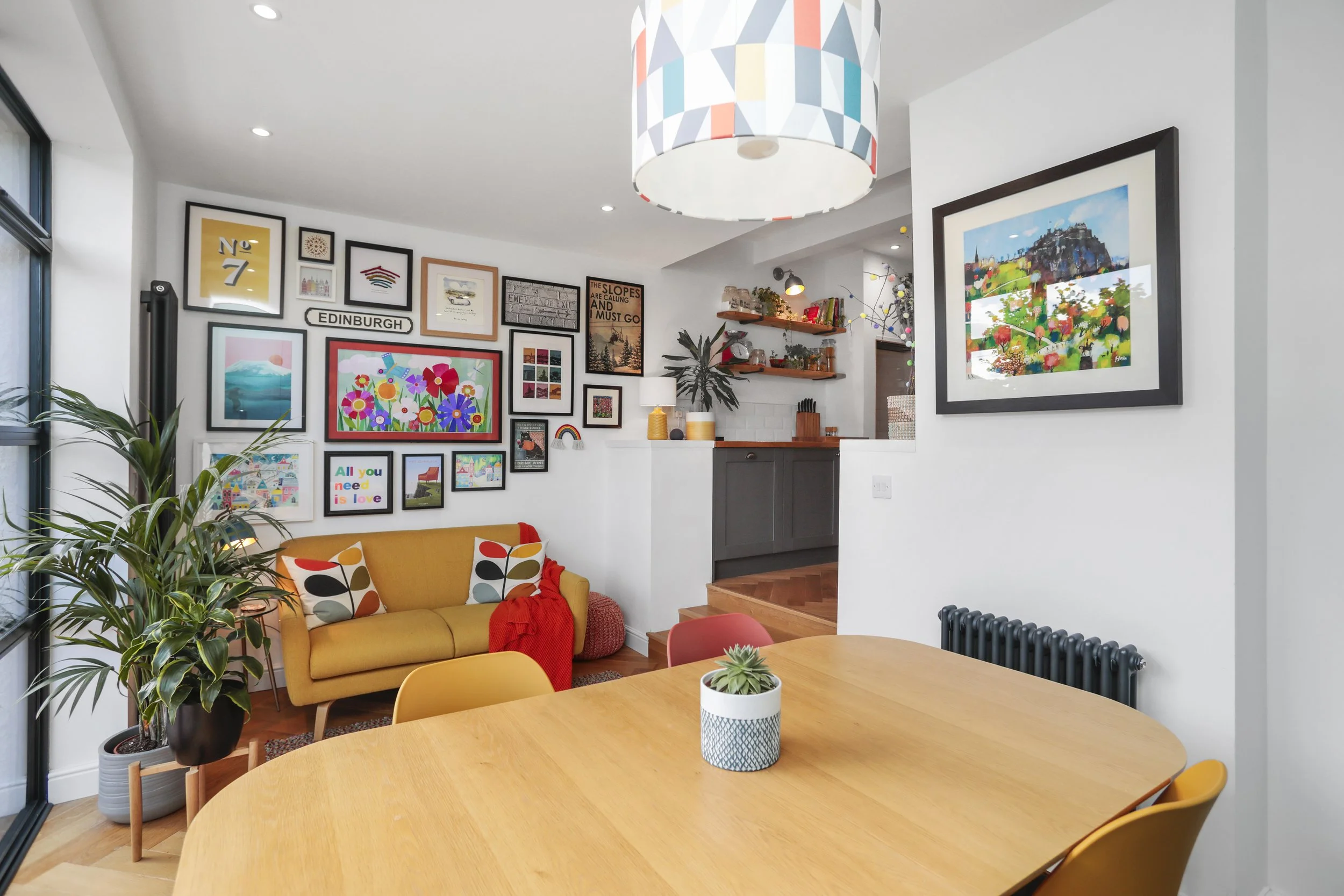 A modern living room and dining area with white walls decorated with framed colorful art prints. There is a yellow couch with patterned pillows, a round wooden dining table with yellow chairs, a potted plant, and a white table centerpiece with a succ