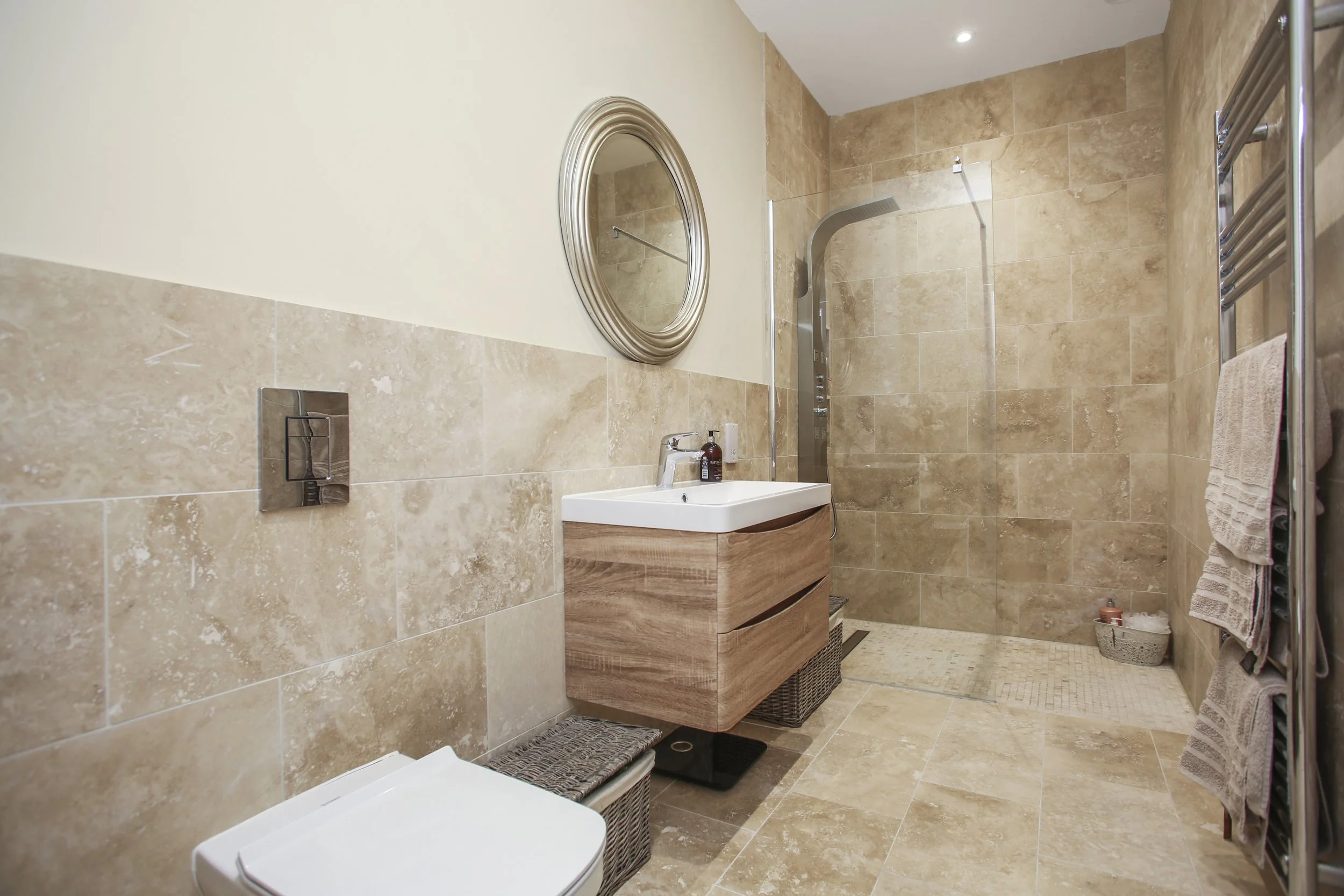 A modern bathroom with beige tile walls and floors, a small sink with a wooden cabinet, a round mirror, a towel rack with beige towels, a shower area with a rain shower head, and a white toilet.