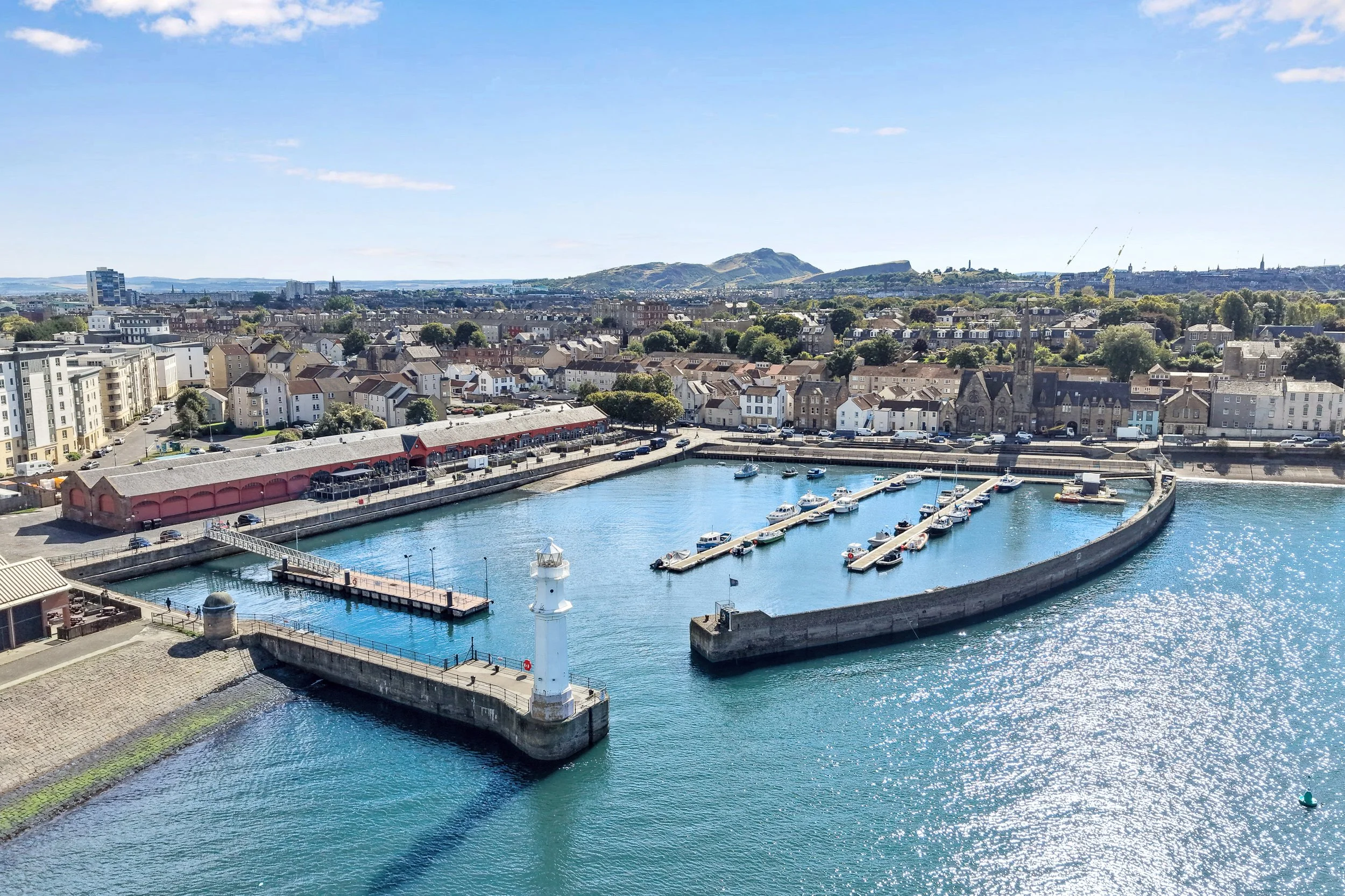 A harbor with boats docked at piers, surrounded by a city with residential buildings, a church with a tall steeple, and green hills in the background on a clear, sunny day.
