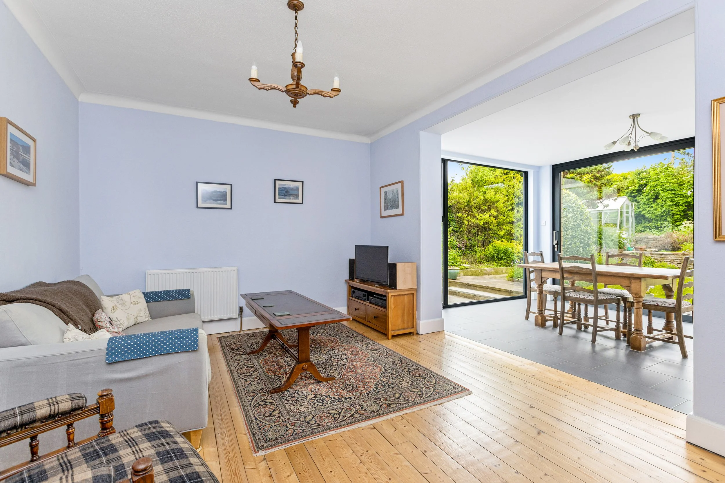 Living room with a sofa, a plaid armchair, a wooden coffee table, and a patterned rug. The room has light blue walls with framed artwork, a small TV on a stand, and large sliding glass doors leading to a garden with greenery and a greenhouse.