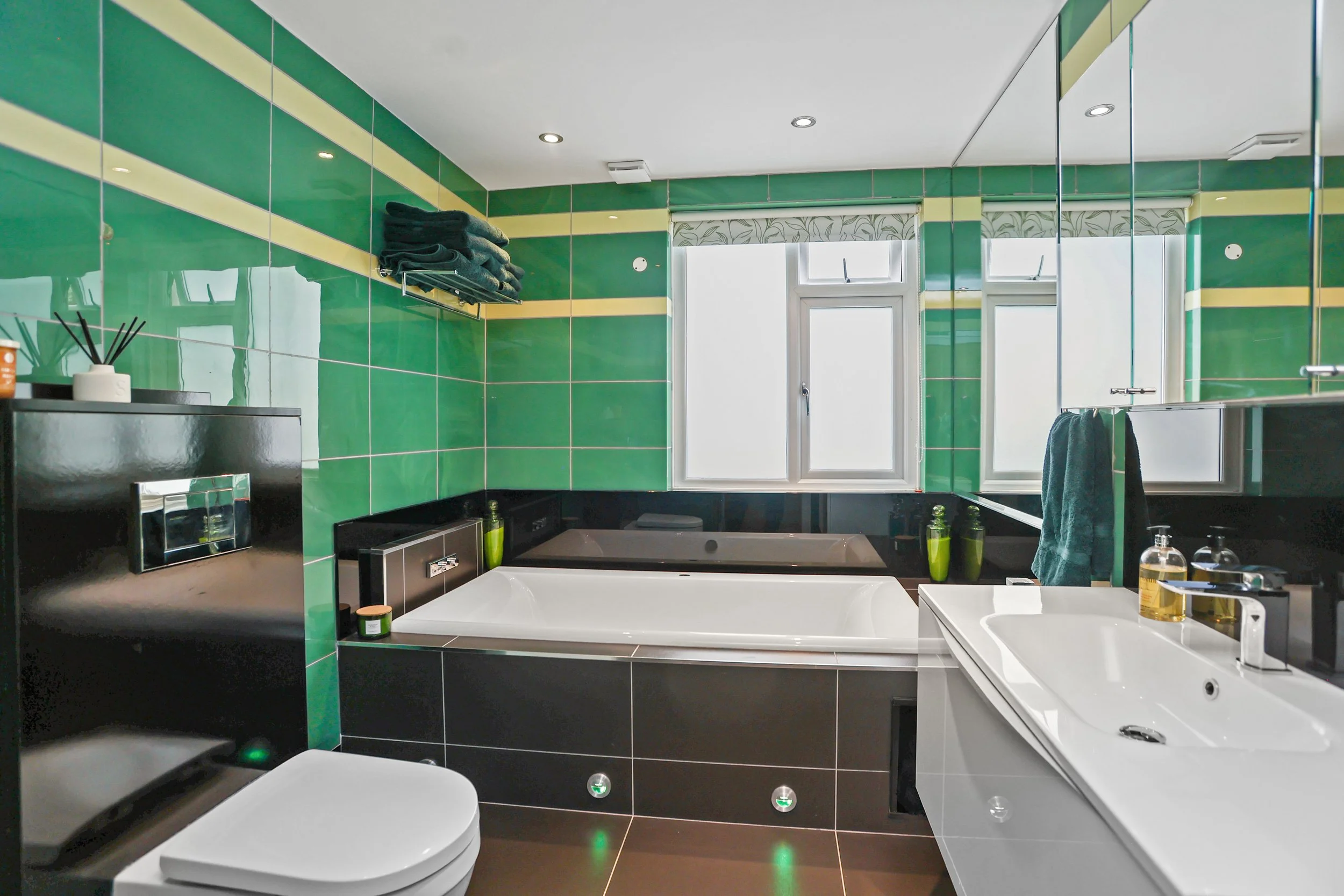 Modern bathroom with dark cabinetry, white bathtub, green and beige striped tiled walls, two windows, and various toiletries on countertop.