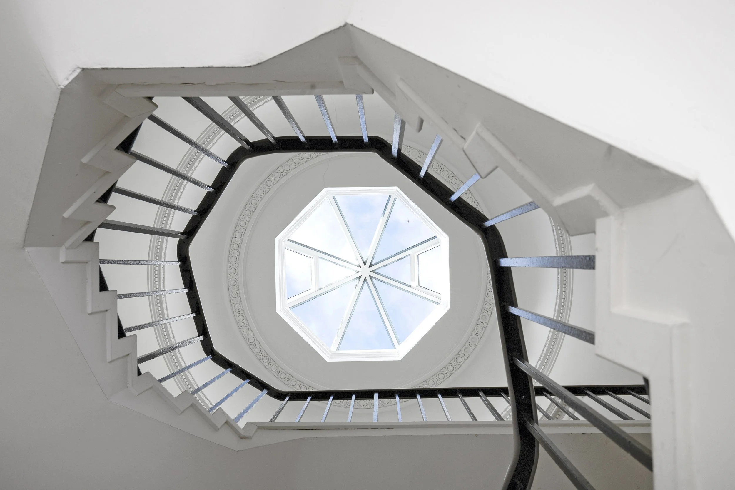 Looking up at a spiral staircase with a large window at the top, showing the sky and some clouds.