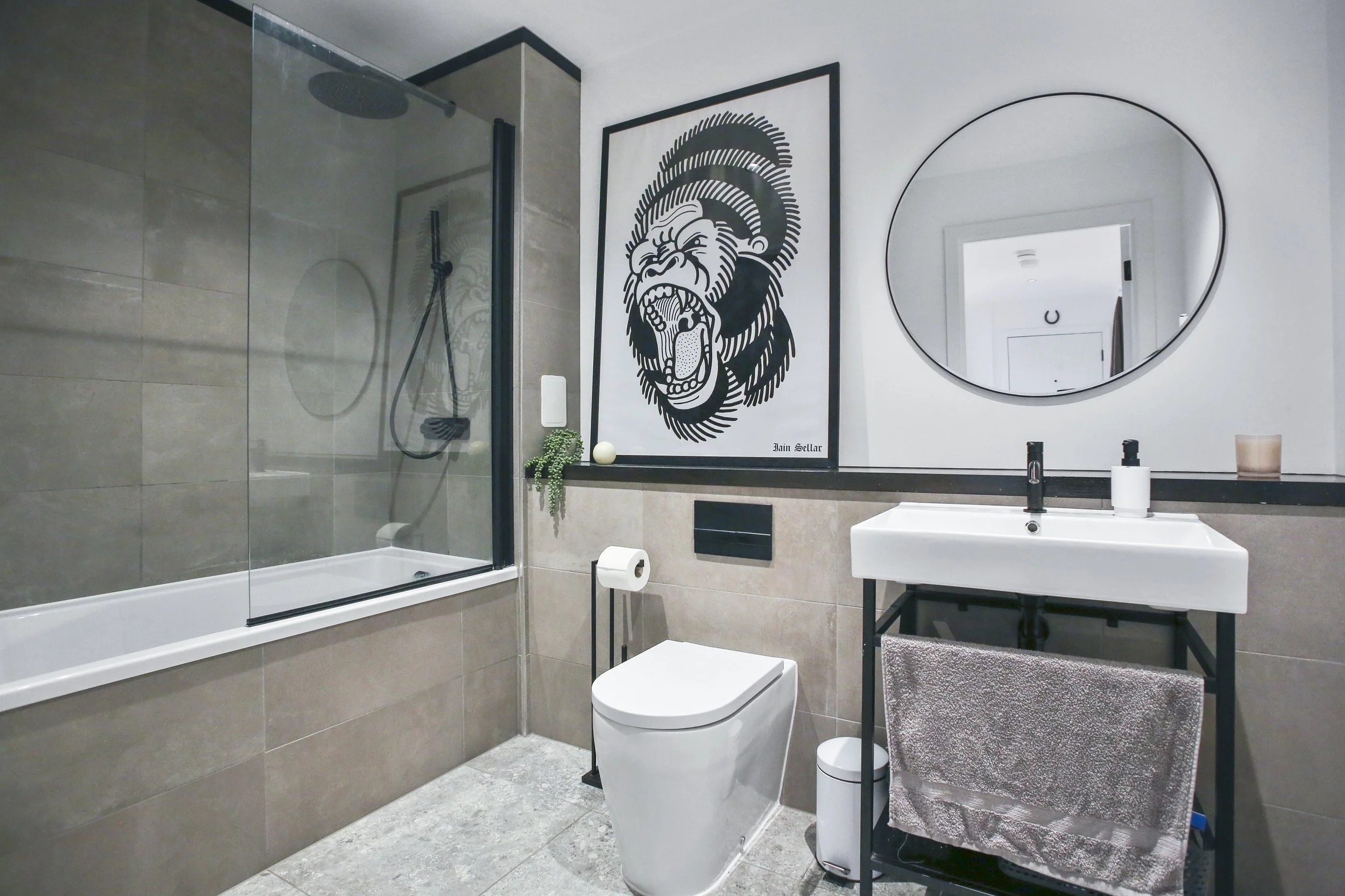 Modern bathroom with a walk-in shower, a framed black and white lion artwork, an oval mirror, a white sink with accessories, and beige tile walls.