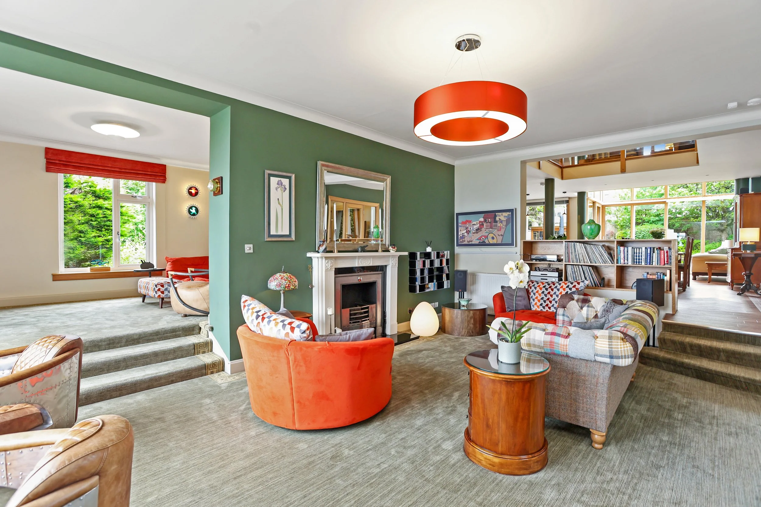 Interior view of a spacious living room with a green accent wall, red and patterned sofas, a white fireplace, and large windows showing greenery outside.