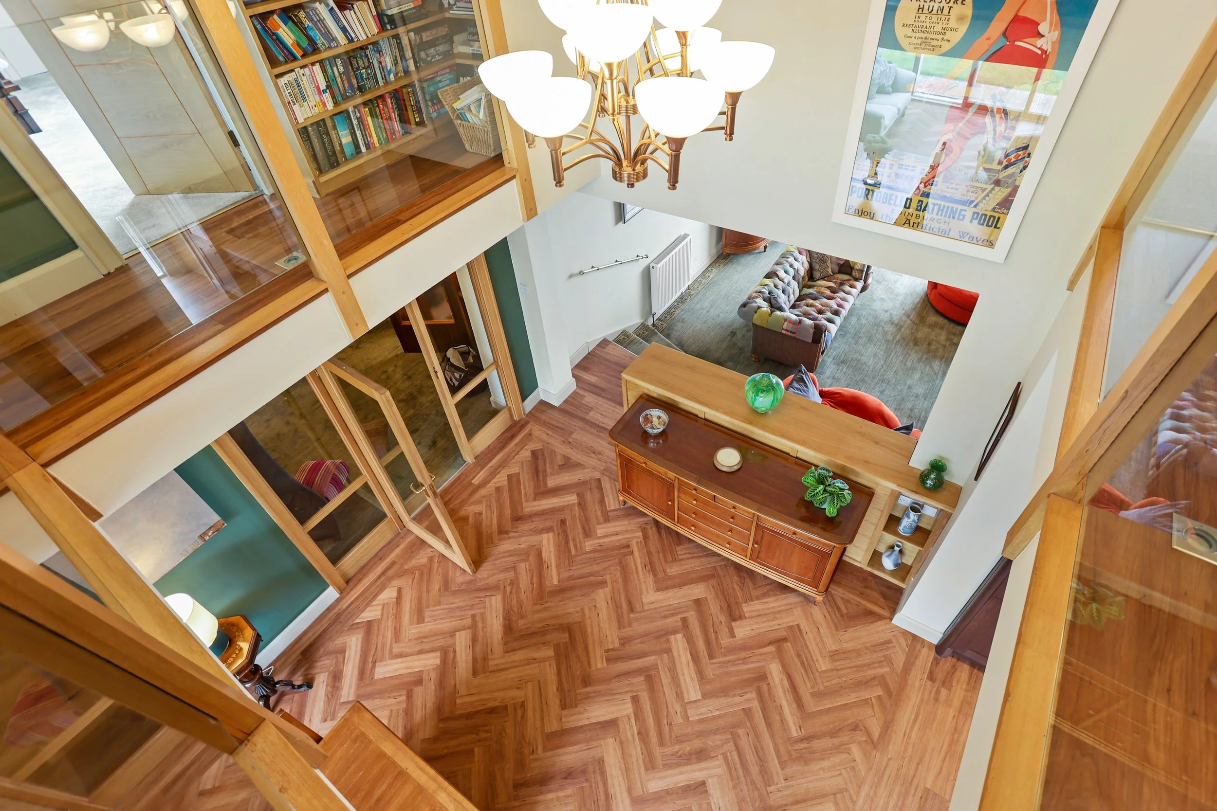 A view from above of a two-story living space with wooden floors, a wooden staircase, a sitting area with a gray couch and colorful pillows, a framed art poster on the wall, a wooden sideboard with decorative items, and a large chandelier with multip