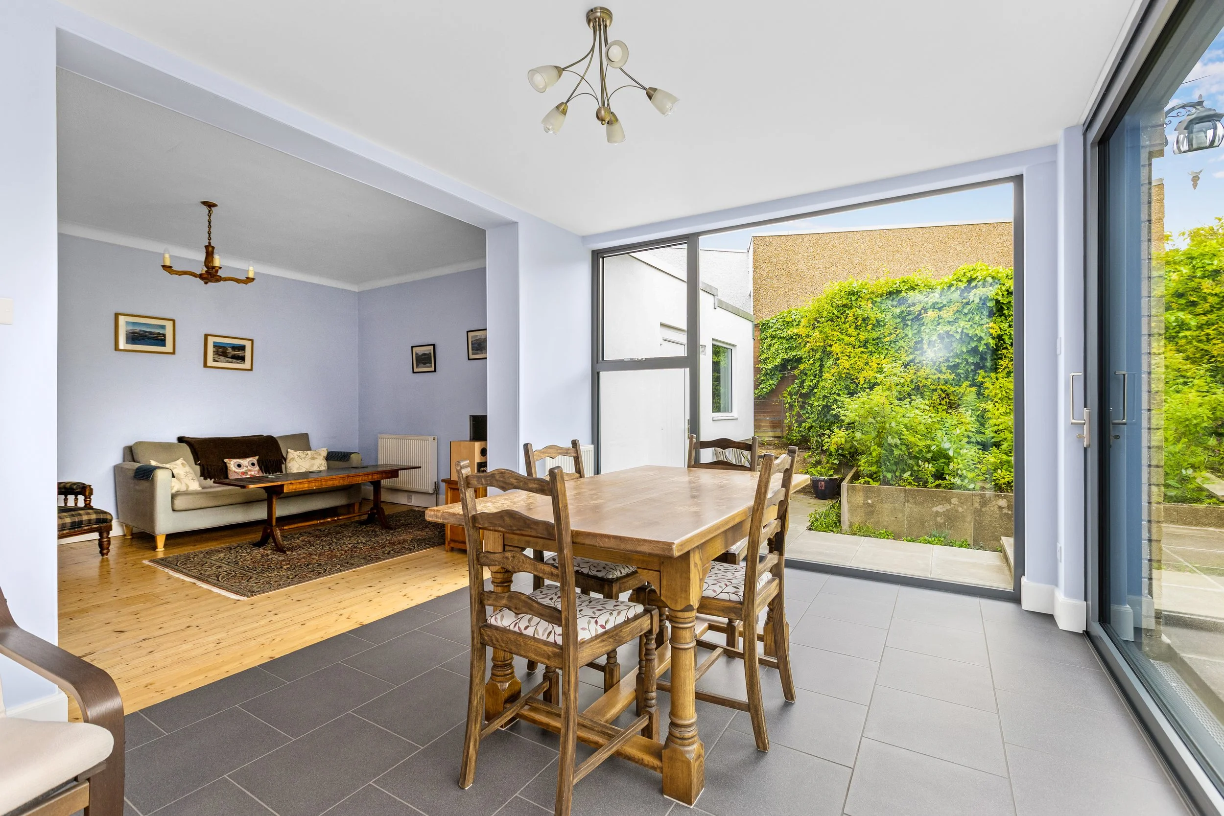 A bright dining and living room area with large glass sliding doors leading to a backyard with green bushes and a concrete patio. The dining table has wooden chairs, and the living room has a couch, framed pictures on the wall, and a chandelier. The 