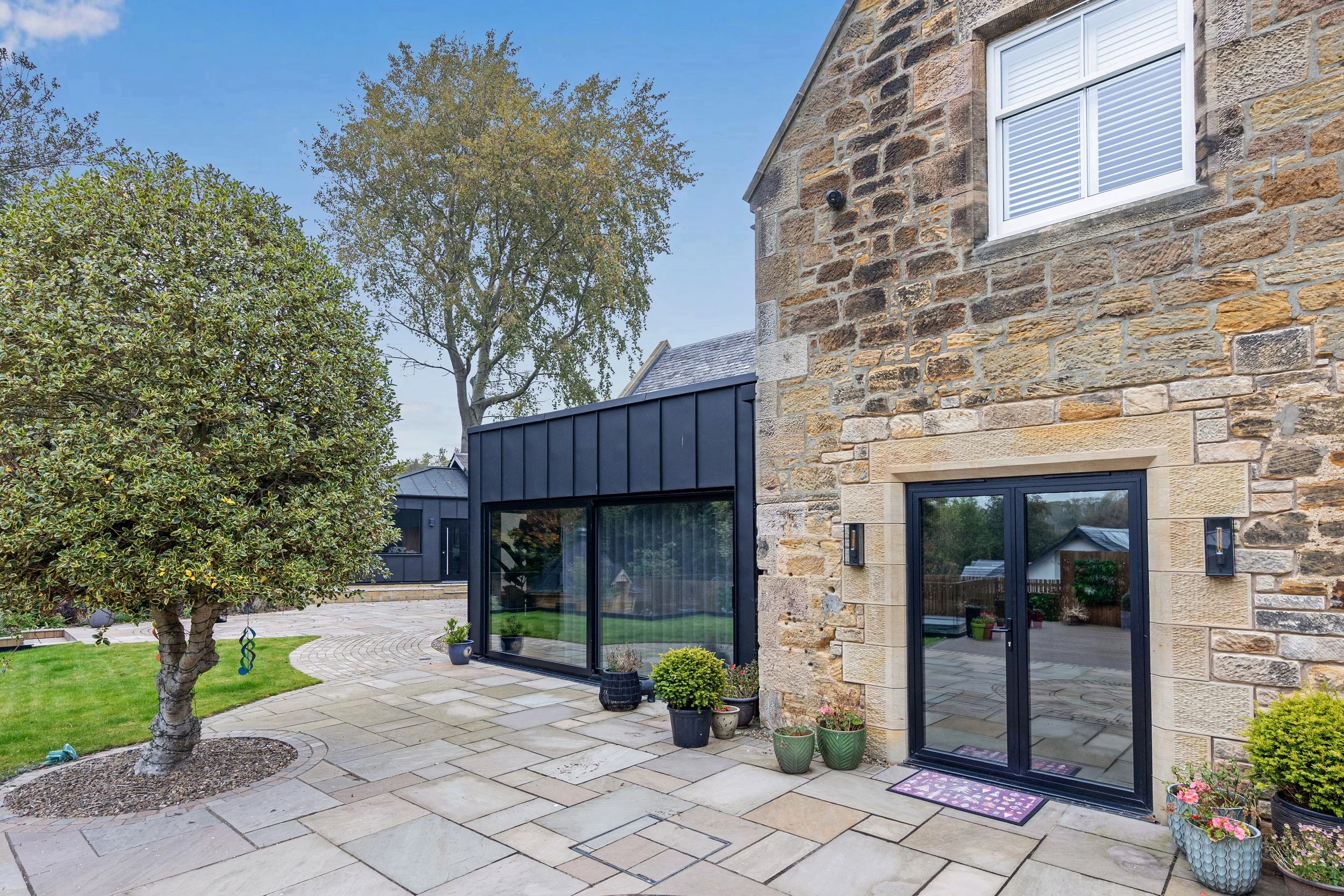 A backyard patio with large stone pavers, potted plants, small trees, a stone house with large windows, and a black fence in the background.