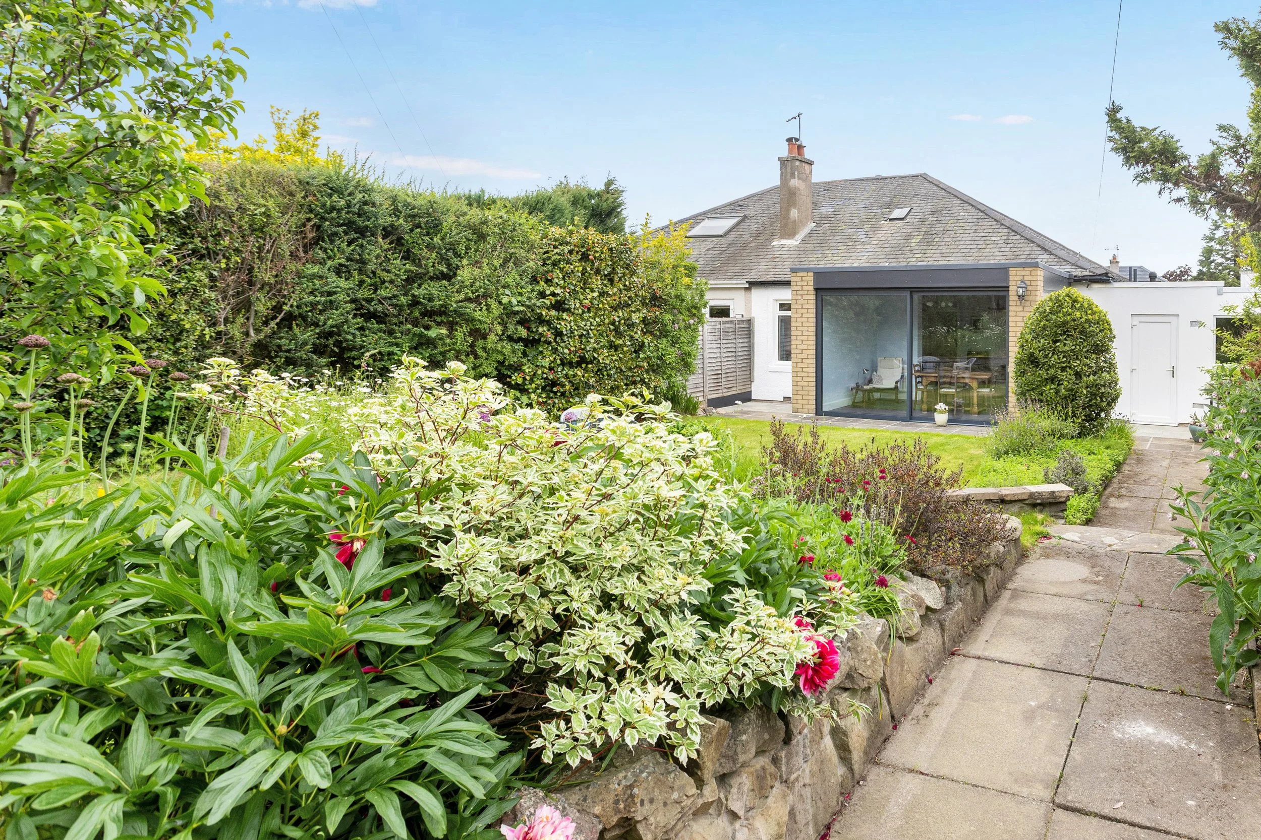 A garden path with green foliage and flowering plants leading to a house with a backyard. The house has a brick and white exterior, a large glass door, and a sloped roof with skylights.