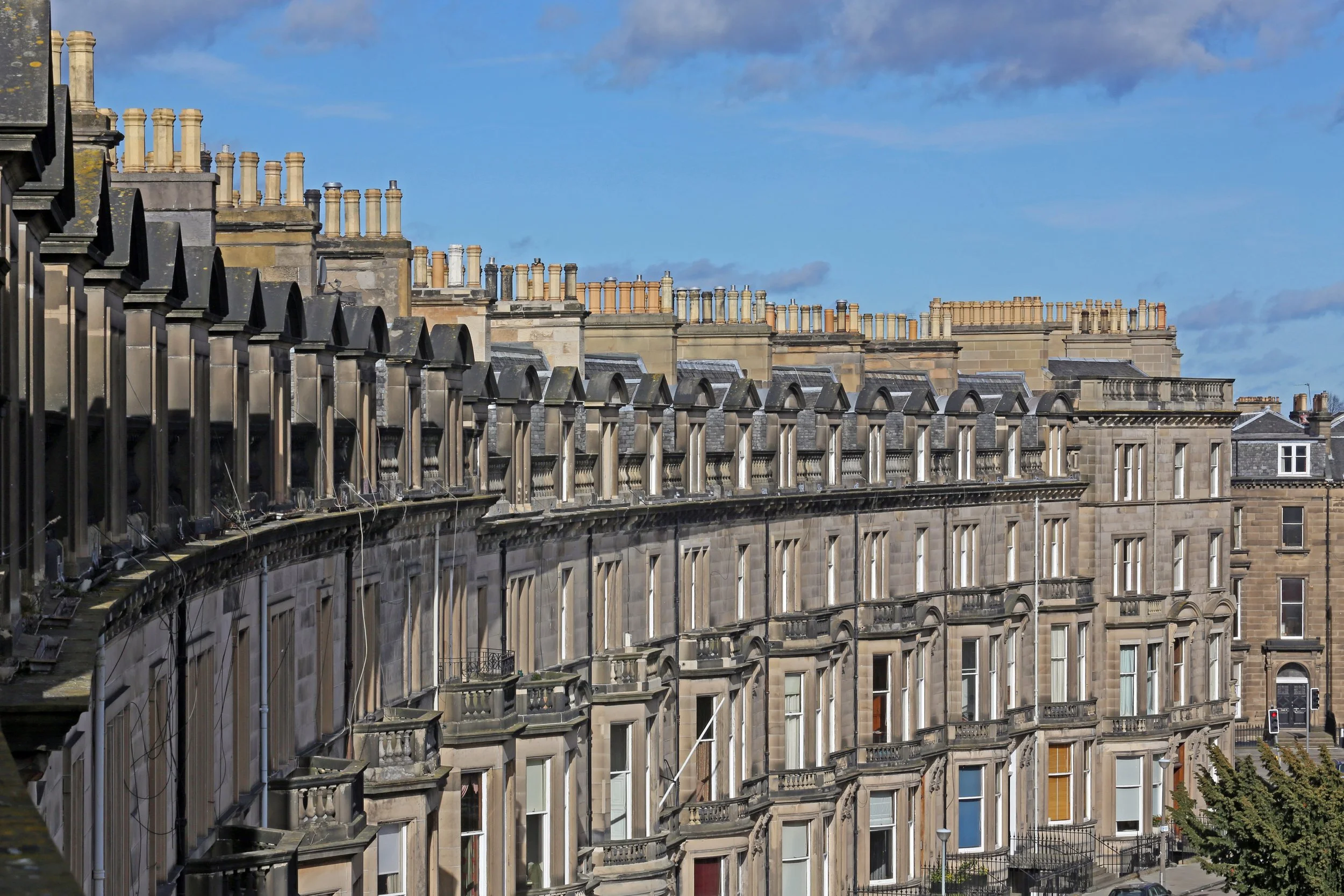 Row of historic stone buildings with multiple chimneys and detailed facades, under a partly cloudy blue sky.