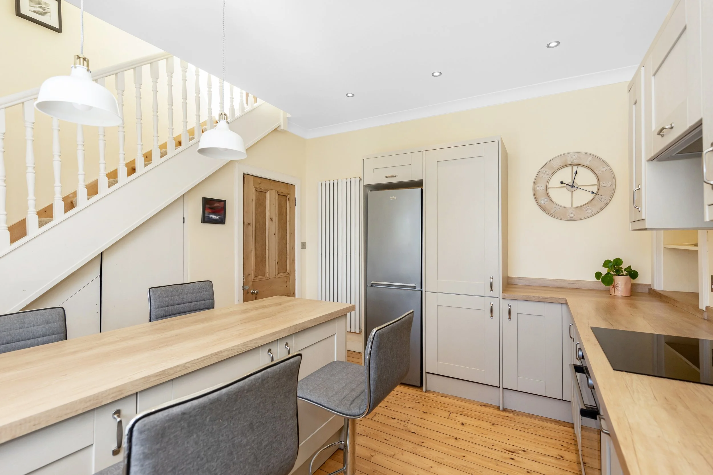 A kitchen with light yellow walls, white cabinets, a wooden dining table with gray chairs, a stainless steel refrigerator, a large wall clock, and a staircase with white spindles and pendant lights.