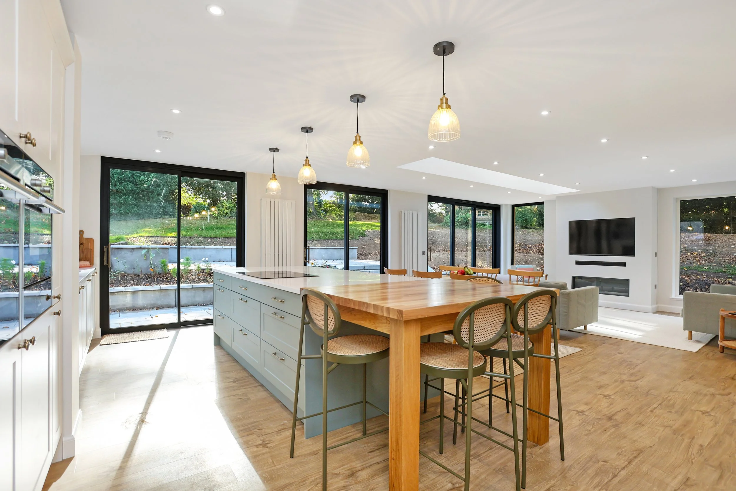Open-concept kitchen and living room with large glass sliding doors leading to a backyard with greenery, pendant lights over a kitchen island, chairs around a wooden dining table, and a television on the wall.