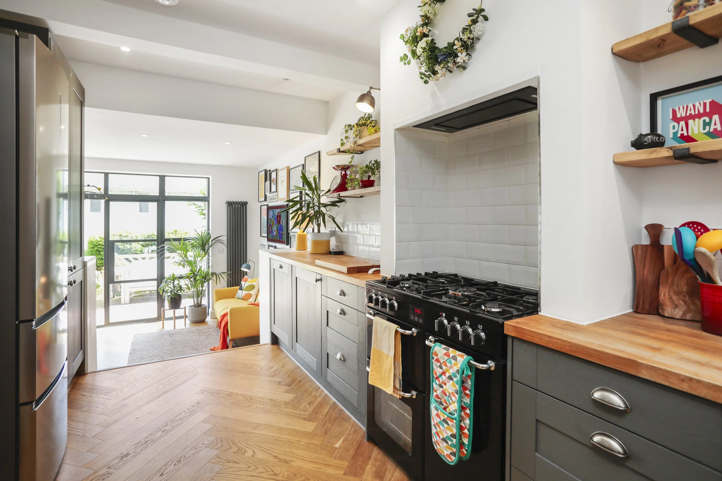 A modern kitchen with gray cabinets, wooden countertops, a black stove, and decorative plants and artwork, with sunlight coming through glass doors in the background.