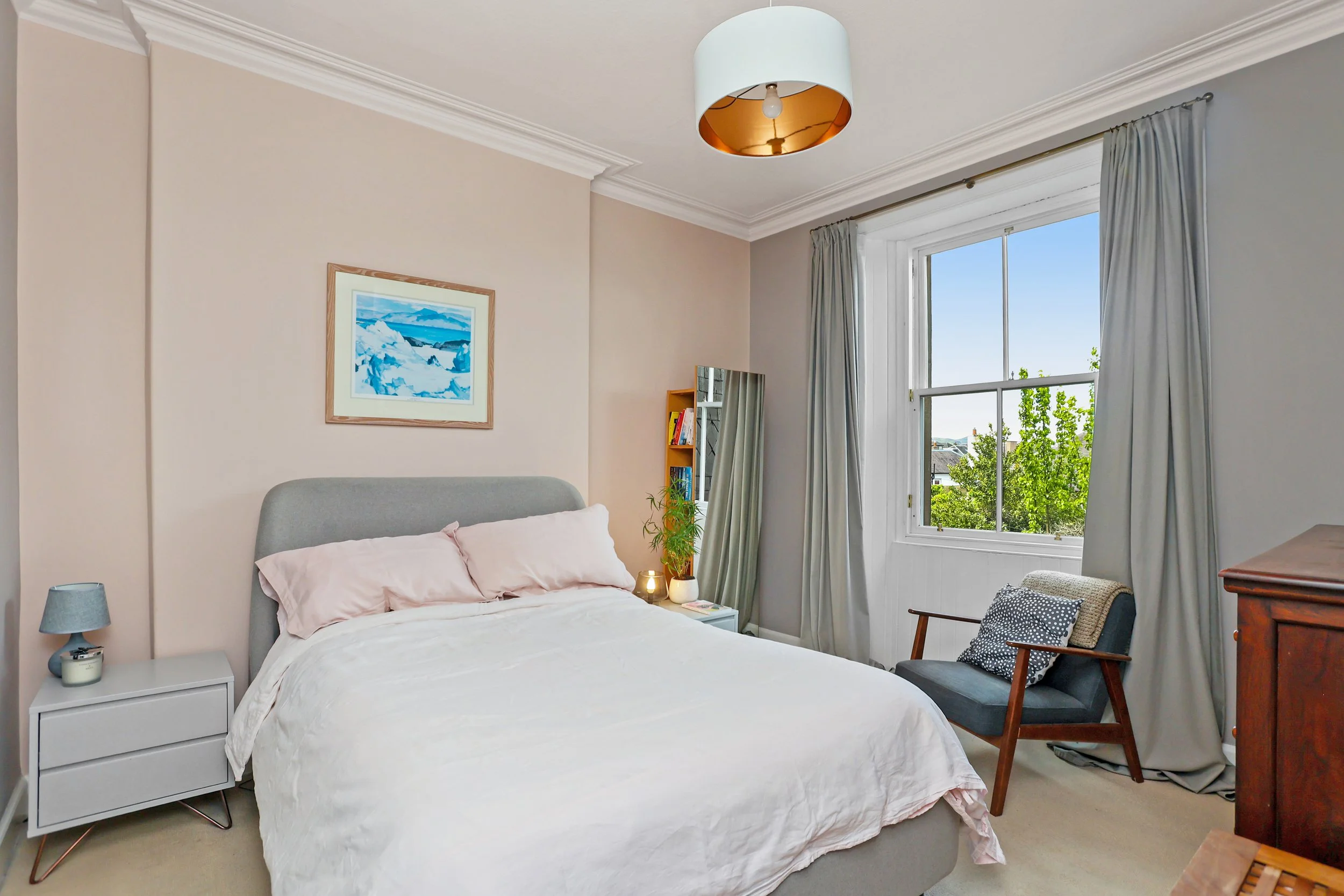 A bedroom with a bed, nightstand, lamp, painting, window with curtains, chair, and wooden dresser.