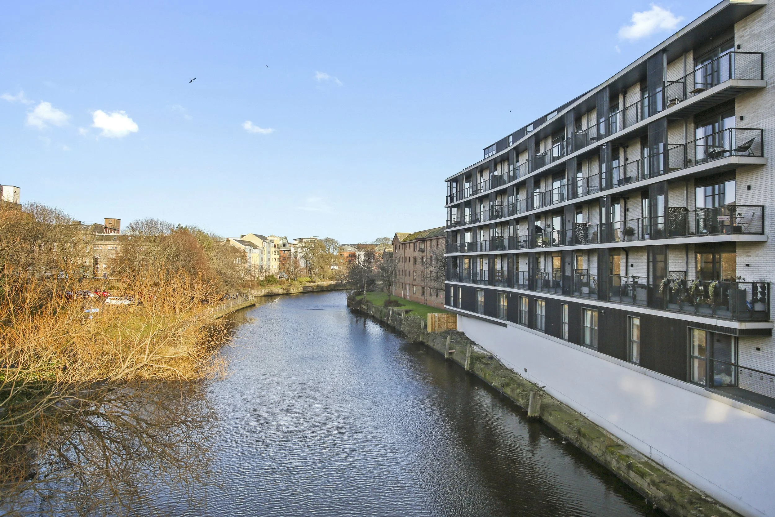 A river with calm water flowing past residential buildings, including a modern apartment complex with balconies, under a clear blue sky with a few clouds and birds flying.