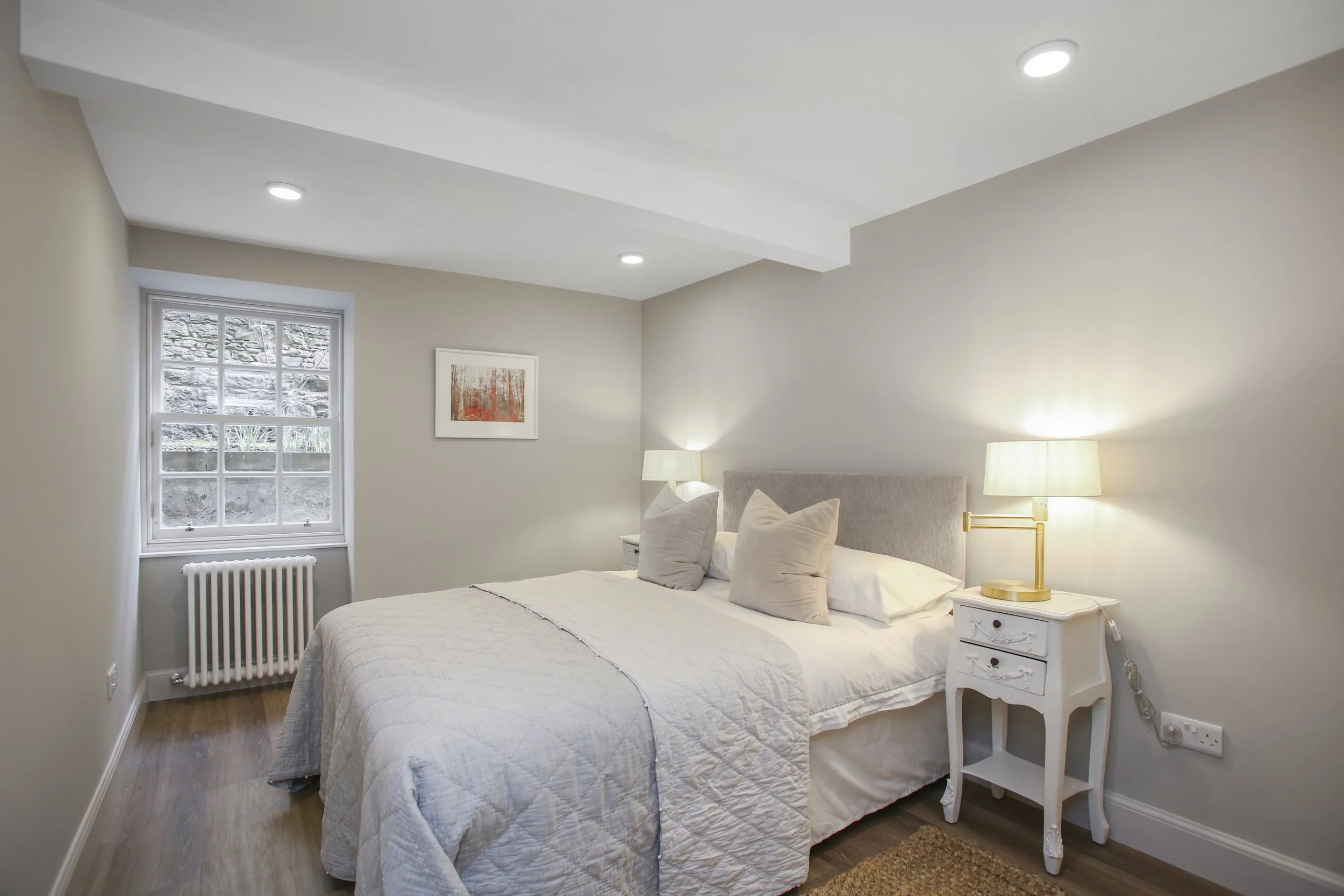 A cozy bedroom with a large bed covered in white quilted bedding and several pillows. On each side of the bed, there are white nightstands, one with a lamp. A window with white framing shows an outdoor scene with rocks and plants. The walls are paint