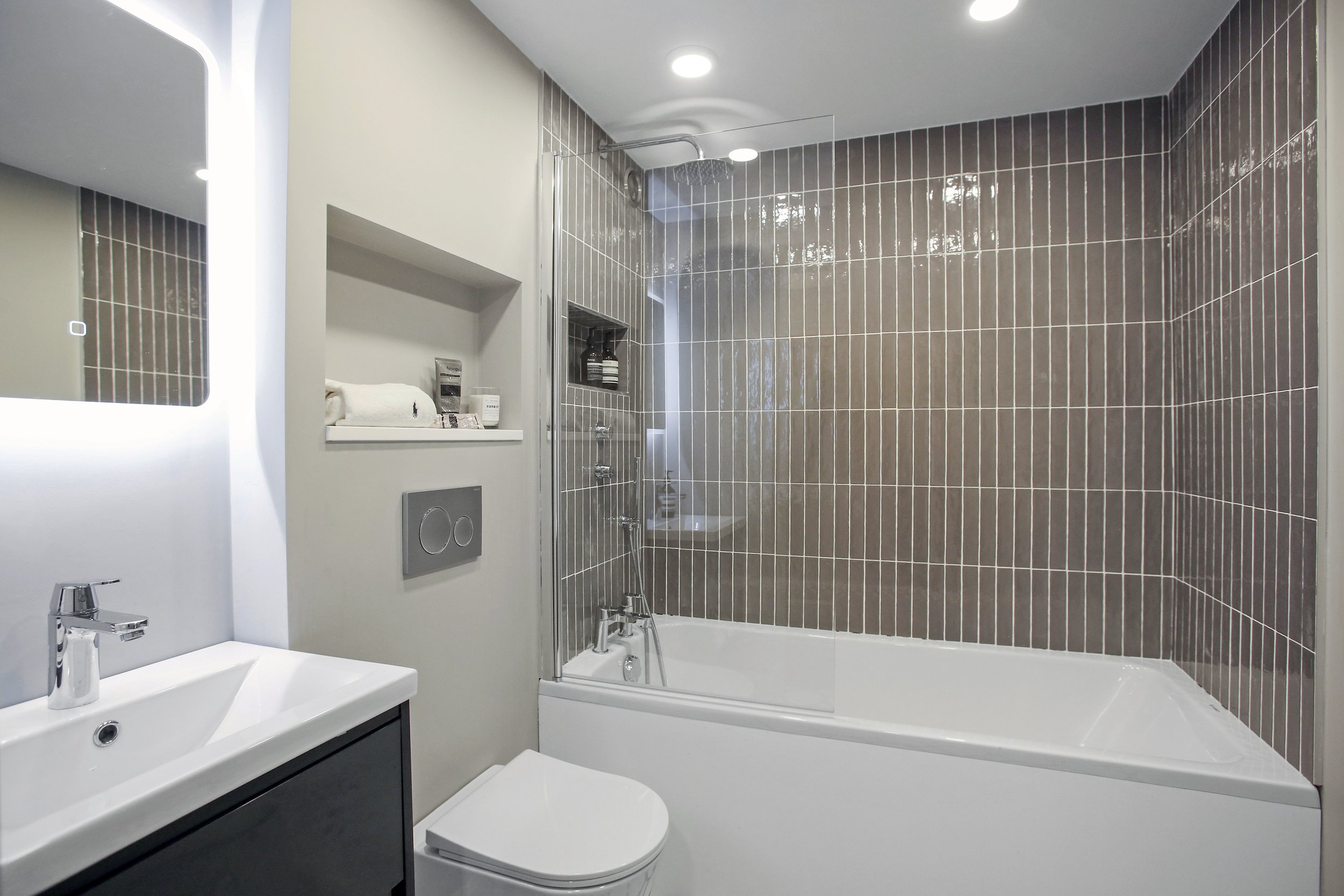 Modern bathroom with a white sink and faucet, a wall-mounted toilet, a bathtub with a glass shower screen, dark brown tiled wall, and a small shelf with toiletries near the shower.