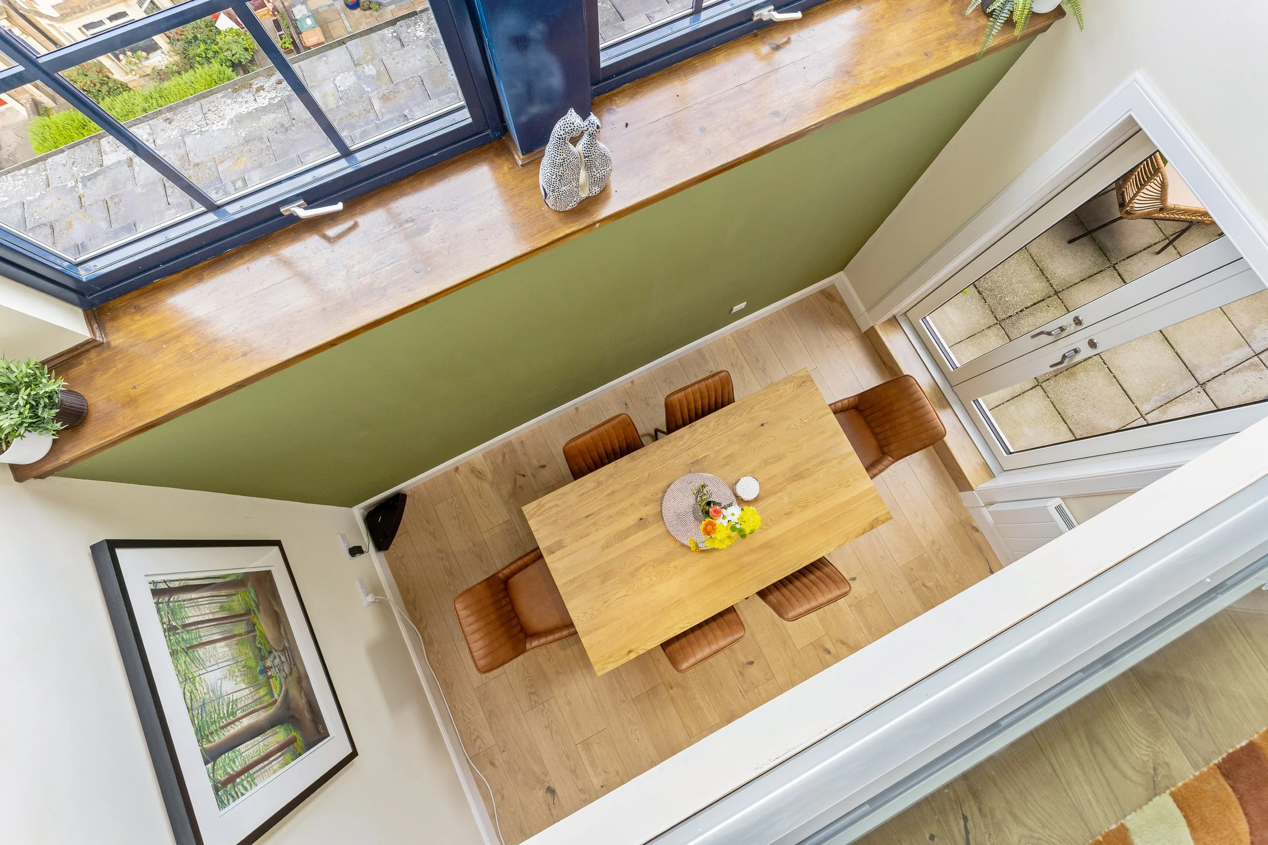 View of a dining area from above, showing a wooden table with a bouquet of yellow, white, and pink flowers, surrounded by four brown leather chairs, large artwork of a tree on the wall, and a window with a wooden ledge displaying a ceramic sculpture 