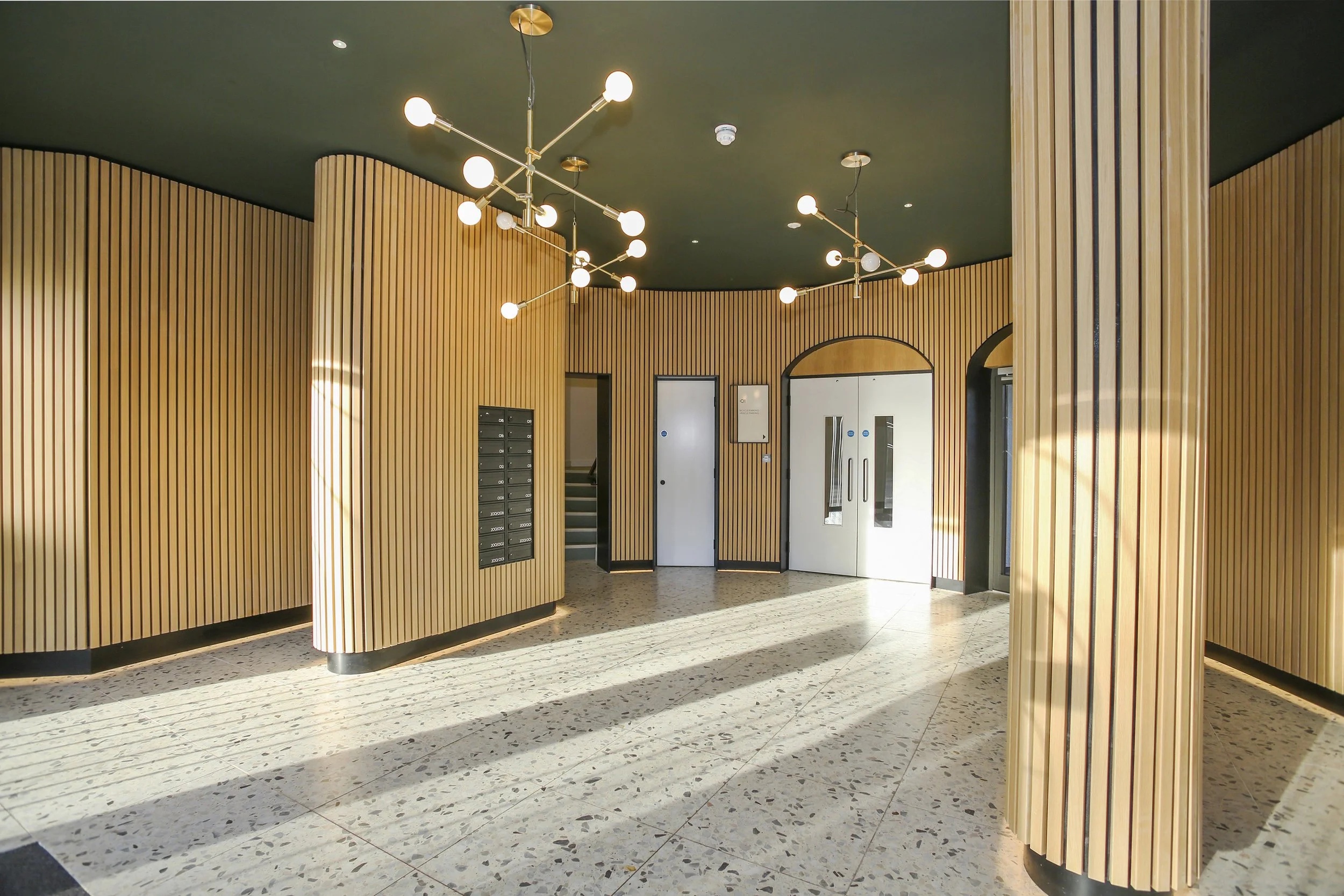Interior of a modern building with wooden panel walls, terrazzo floors, white double doors, and unique gold and white lighting fixtures on a green ceiling.