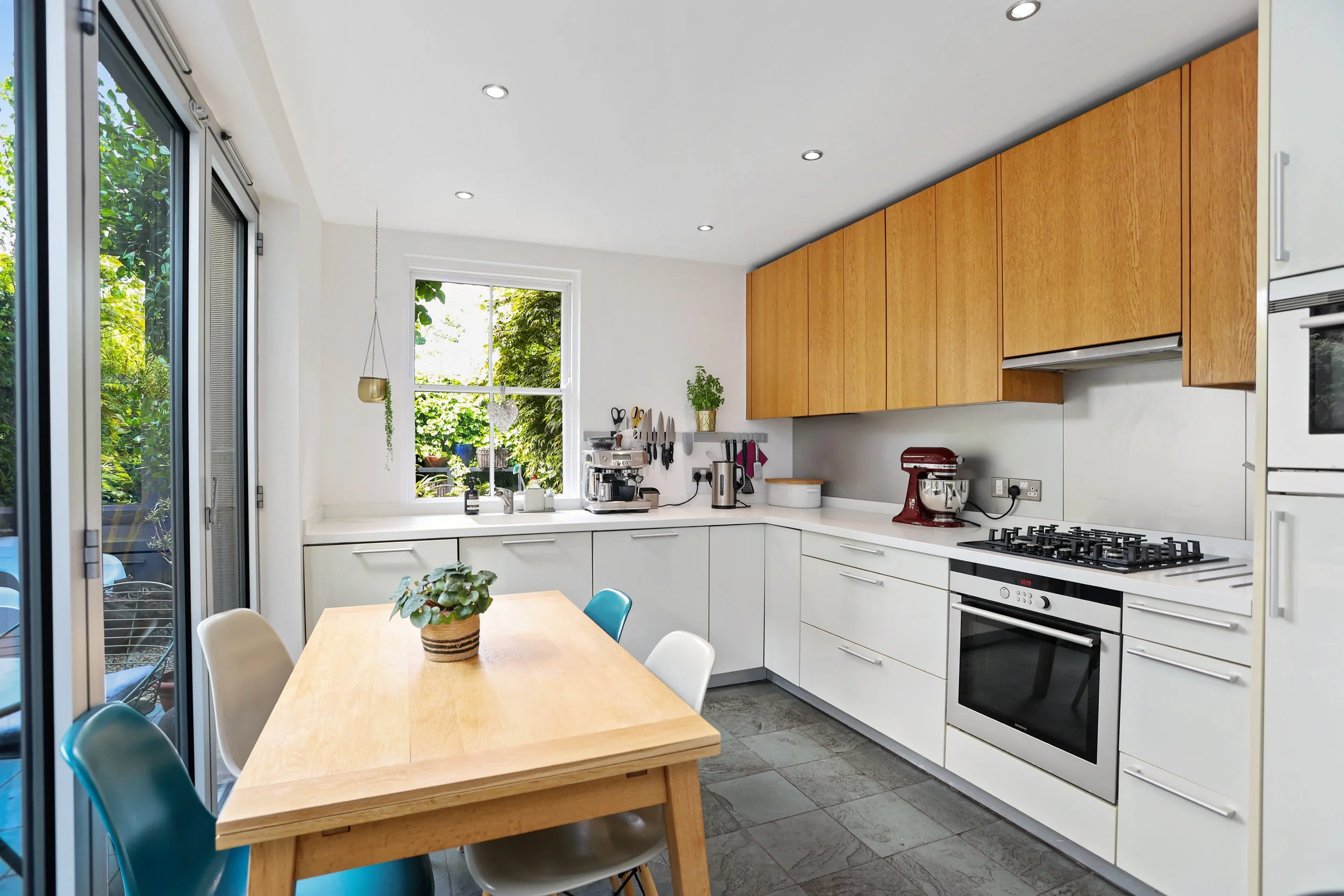 A bright, modern kitchen with white cabinets, wooden upper cabinets, and a white countertop. There are two small windows overlooking a green outdoor garden. A wooden dining table with colorful chairs is in the center. The kitchen features a gas stove
