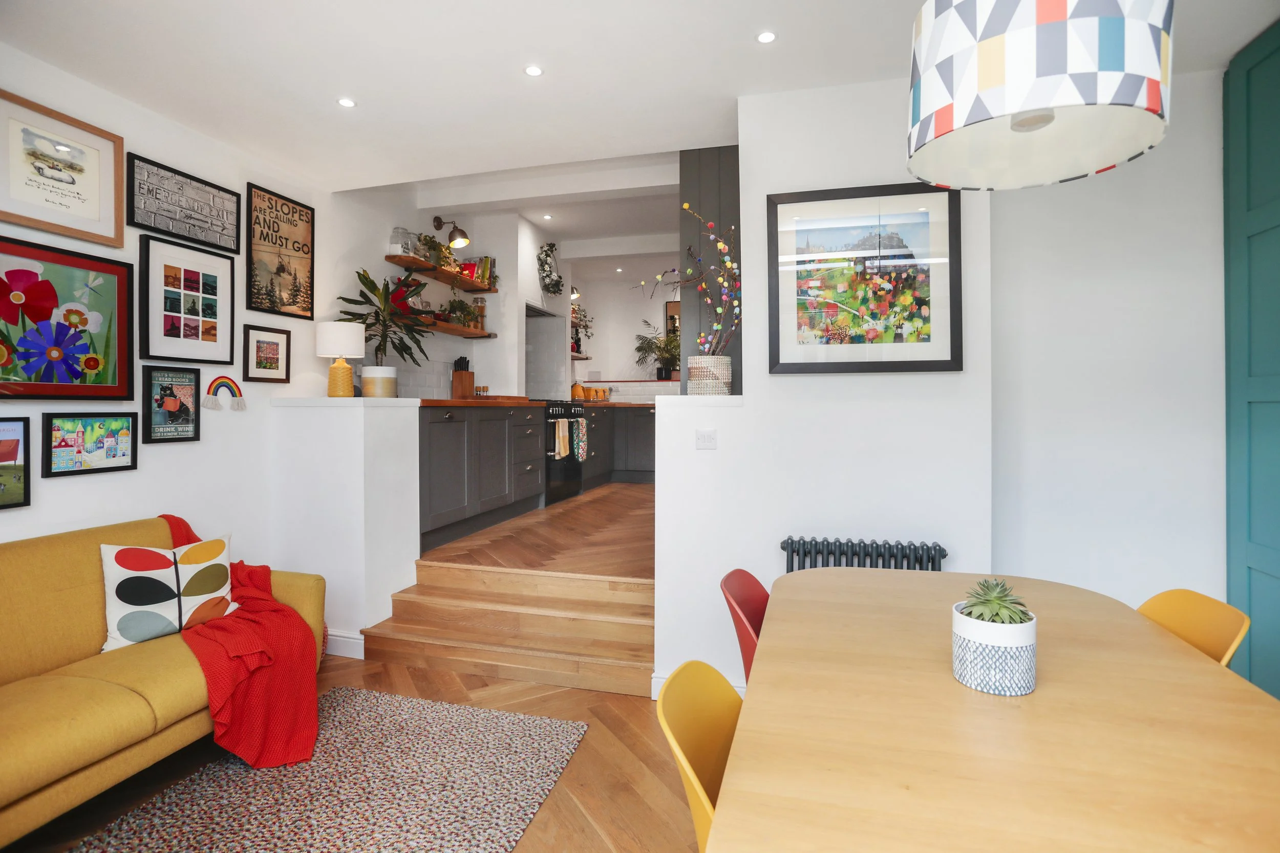 Living room with yellow sofa, colorful pillows, red throw blanket, gallery wall with framed art, wooden dining table with yellow chairs, potted succulent, and a view of the kitchen with dark cabinets and wall art